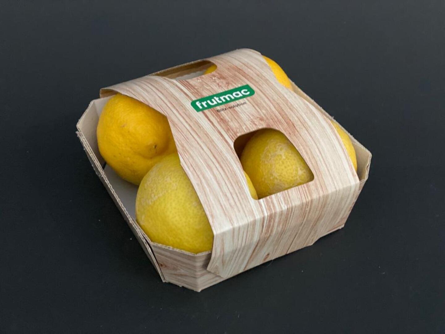 Ein Kartonbehälter mit vier gelben Zitronen darin, teilweise von einer Hülle mit Holzmaserung bedeckt, auf der „frutmac“ steht, vor einem schlichten dunklen Hintergrund.