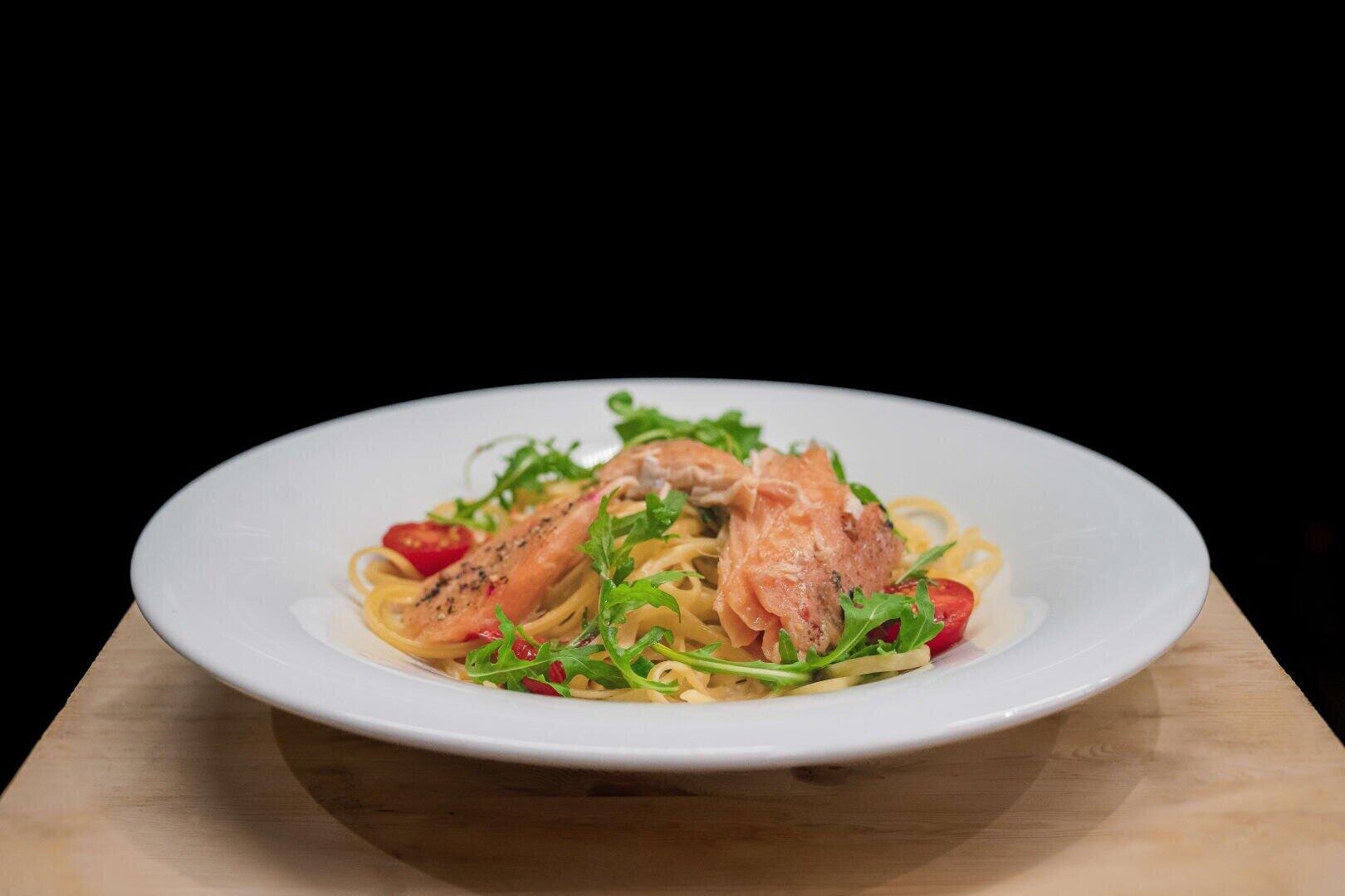 Eine weiße Schüssel Pasta, garniert mit Lachs, Rucola und halbierten Kirschtomaten.
