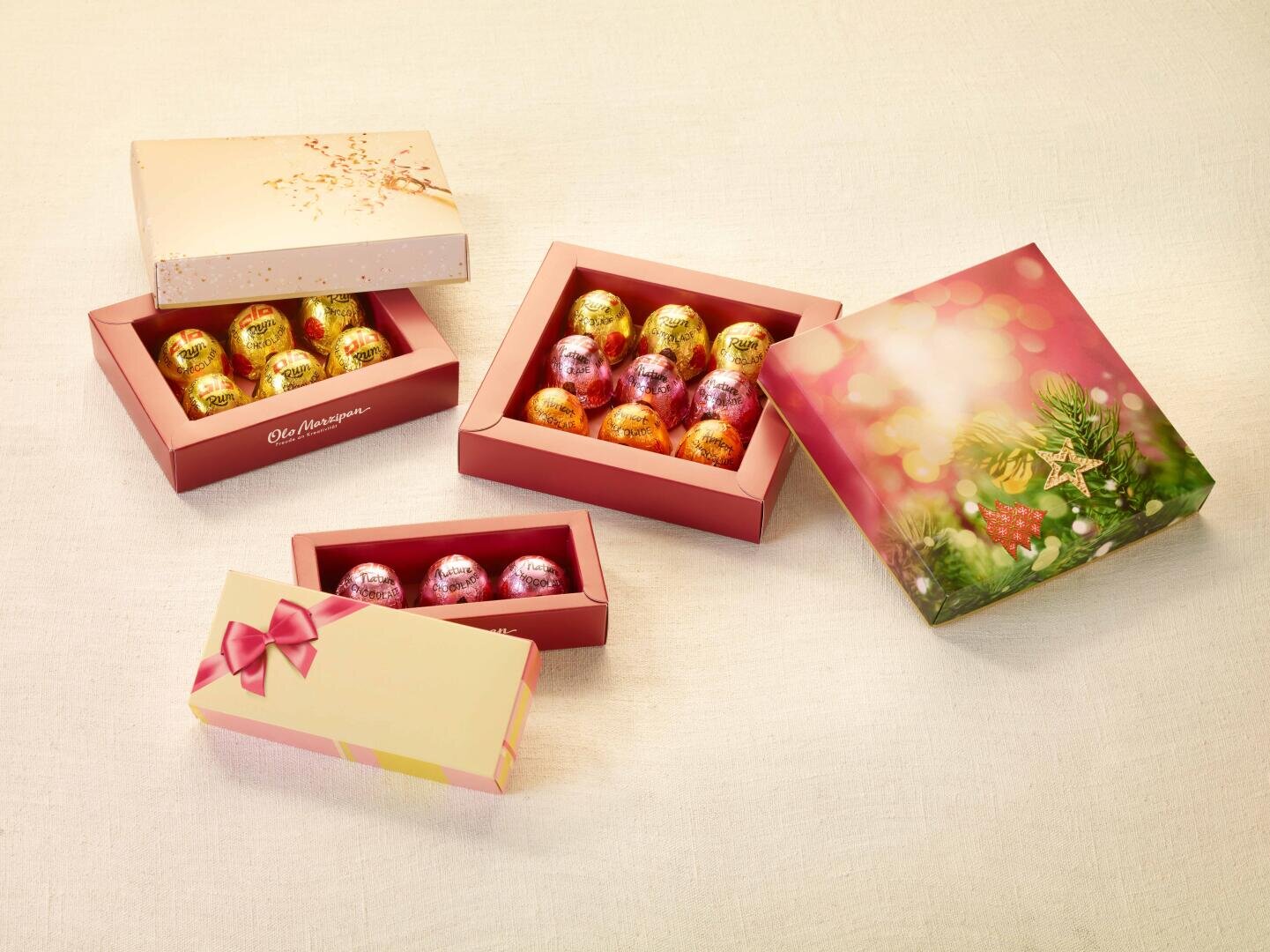 Vier festliche Schachteln, von denen zwei geöffnet sind und in denen in Folie verpackte Schokoladeneier in Gold, Rosa, Orange und Rot zum Vorschein kommen. Die Schachteln sind mit weihnachtlichen Motiven verziert und stehen auf einer hellen, strukturierten Unterlage.