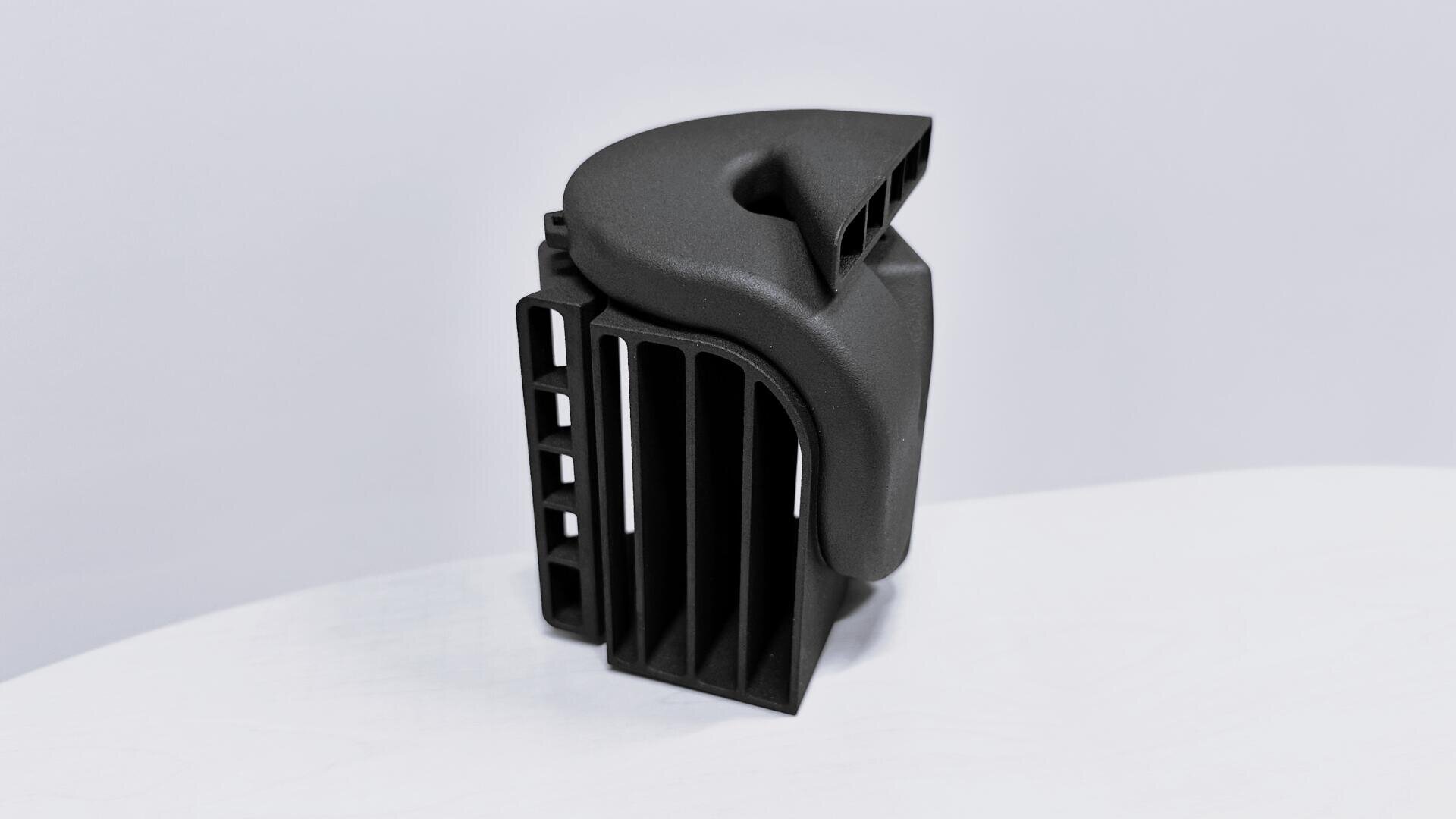 Ein schwarzes, im 3D-Druckverfahren hergestelltes Kunststoffbauteil mit geschwungenen und rechteckigen Abschnitten liegt auf einer weißen Fläche vor einem hellgrauen Hintergrund.