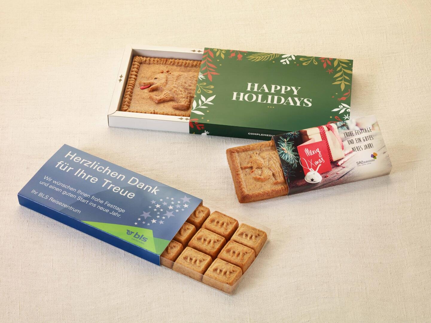 Drei festliche Plätzchenschachteln: Auf einer steht „HAPPY HOLIDAYS“ und darauf ist ein Rentierplätzchen abgebildet, auf einer anderen steht „Herzlichen Dank für Ihre Treue“ und darin befinden sich quadratische Plätzchen, und die dritte Schachtel ist mit einem Weihnachtsgruß und einem Stern-Ornament verziert.