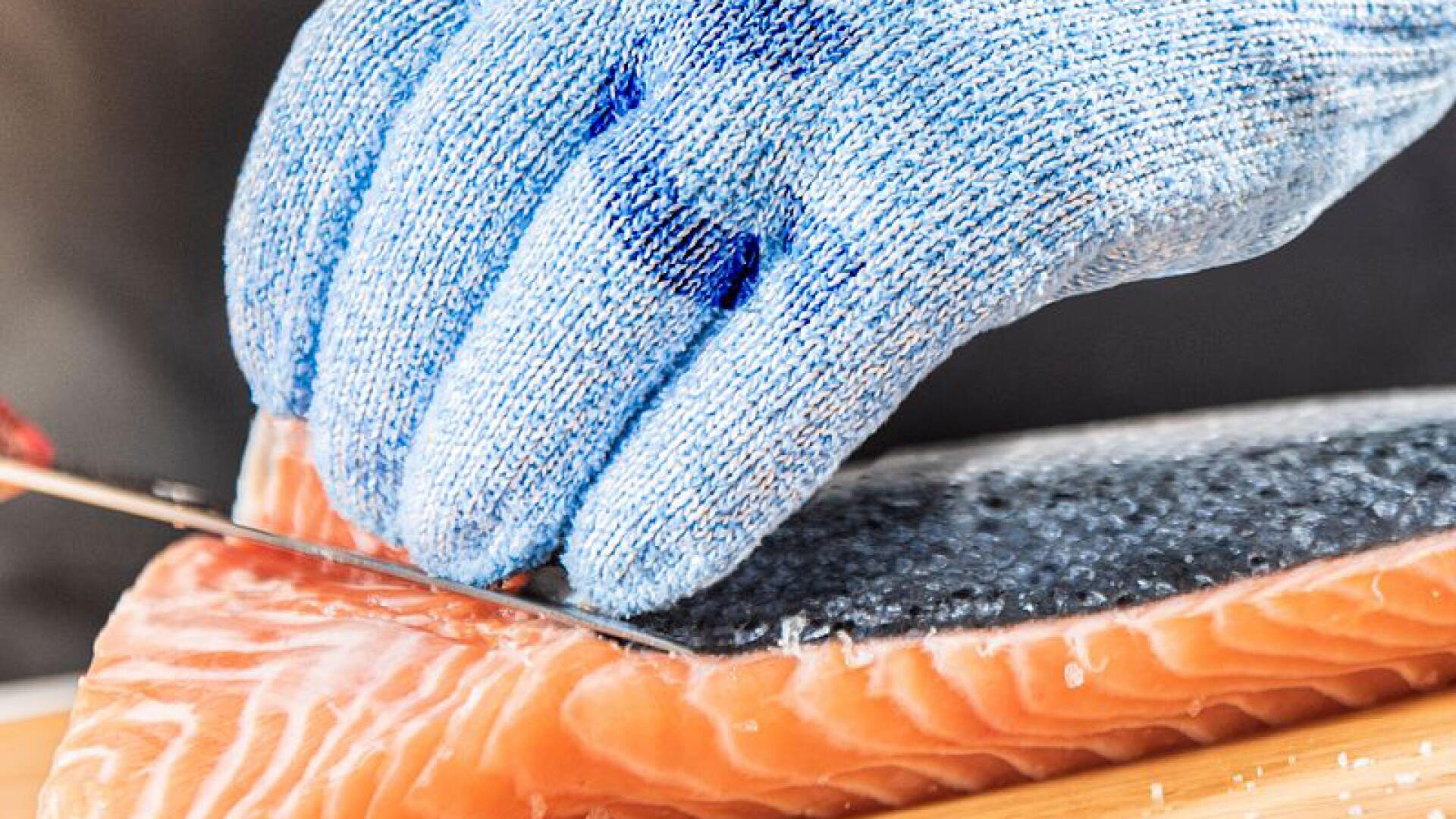Eine Person, die einen blauen, schnittfesten Handschuh trägt, entfernt mit einem Messer die Haut von einem frischen Lachsfilet auf einem Holzschneidebrett.