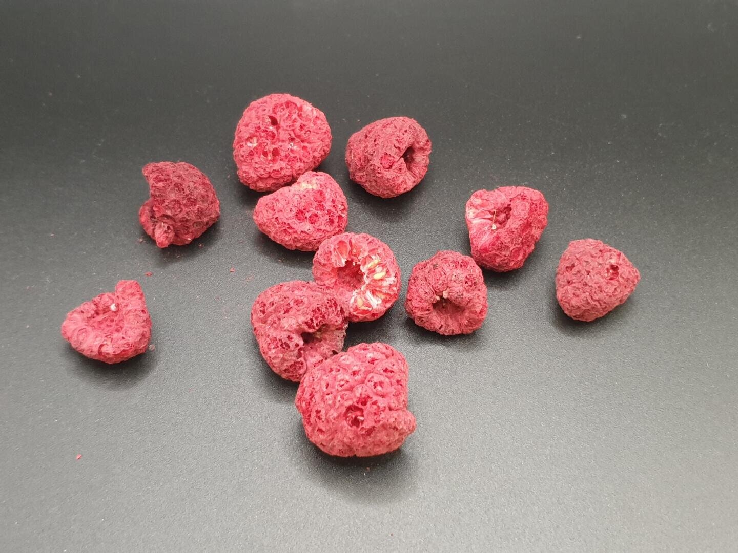 Auf einer glatten, dunklen Oberfläche liegen mehrere gefriergetrocknete Himbeeren verstreut. Die Beeren sind leicht, knusprig und haben eine runzlige Oberfläche sowie eine leuchtend rosa-rote Farbe.
