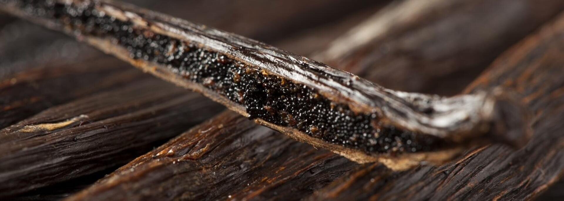 Eine Nahaufnahme einer aufgeschnittenen Vanilleschote, die die winzigen schwarzen Samen und das feuchte Innere zeigt, während im Hintergrund andere ganze Vanilleschoten unscharf zu sehen sind.