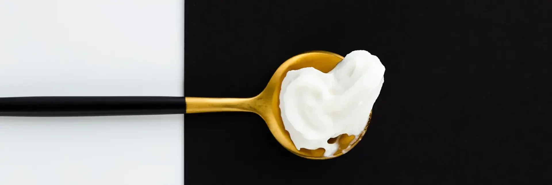 Auf einem Hintergrund, der zu gleichen Teilen schwarz und weiß ist, liegt ein Klecks weißer Sahne oder Joghurt auf einem goldenen Löffel mit schwarzem Stiel.
