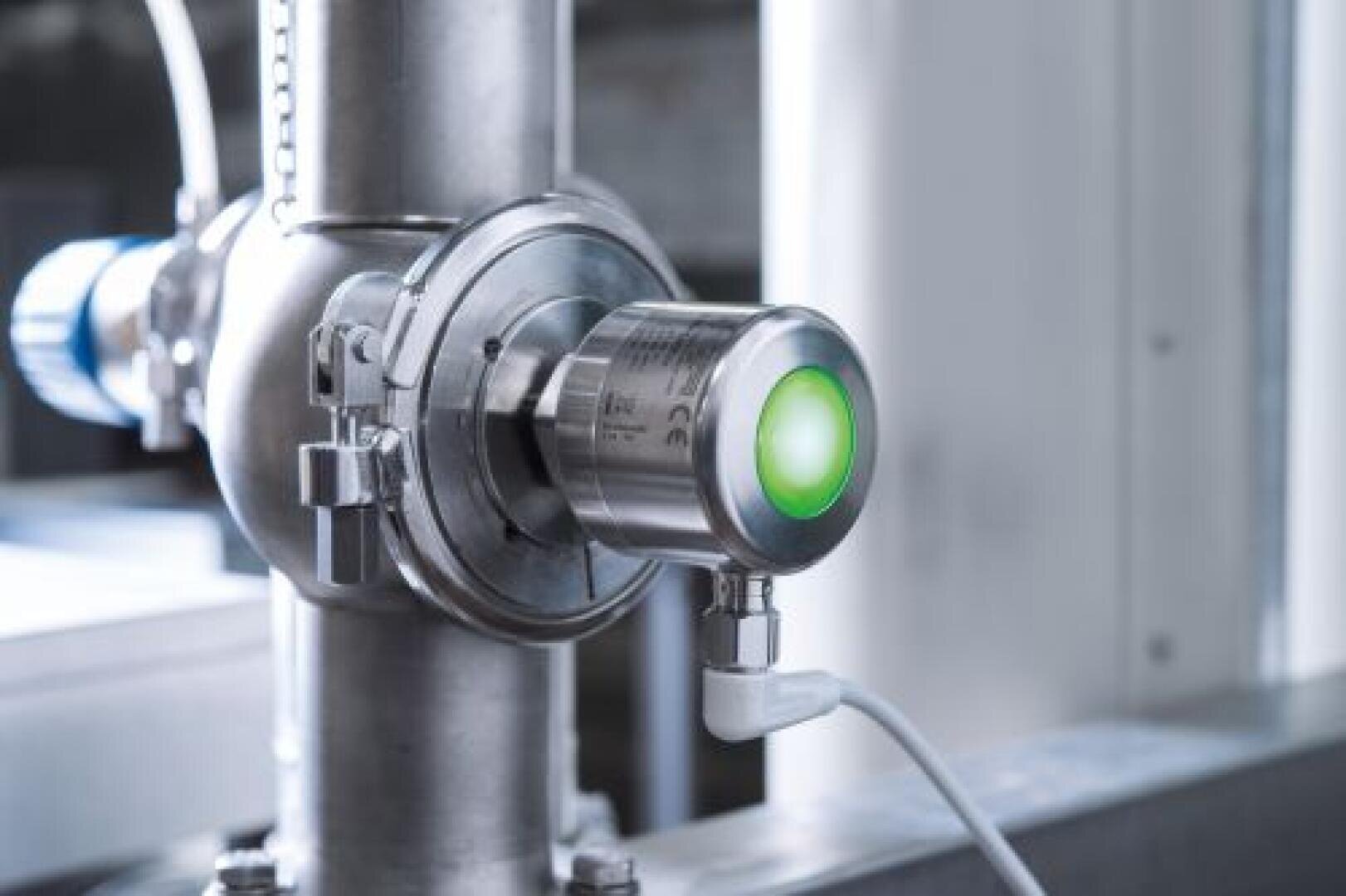 Ein metallener Industriesensor mit einem grün leuchtenden Licht ist mit einer Klemme an einem Rohr befestigt. Ein weißes Kabel ist an den Sensor angeschlossen, und der Hintergrund ist unscharf, sodass ein Teil der industriellen Umgebung zu sehen ist.