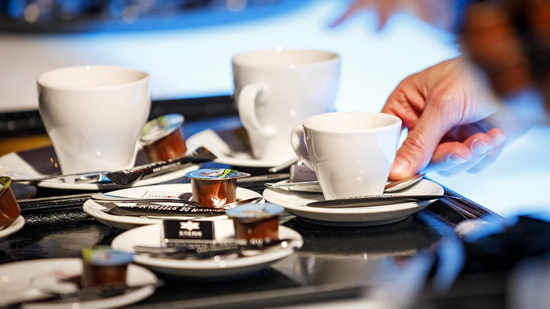 Eine Hand stellt eine kleine weiße Espressotasse auf ein Tablett, auf dem bereits Kaffeetassen und Milchkännchen stehen.