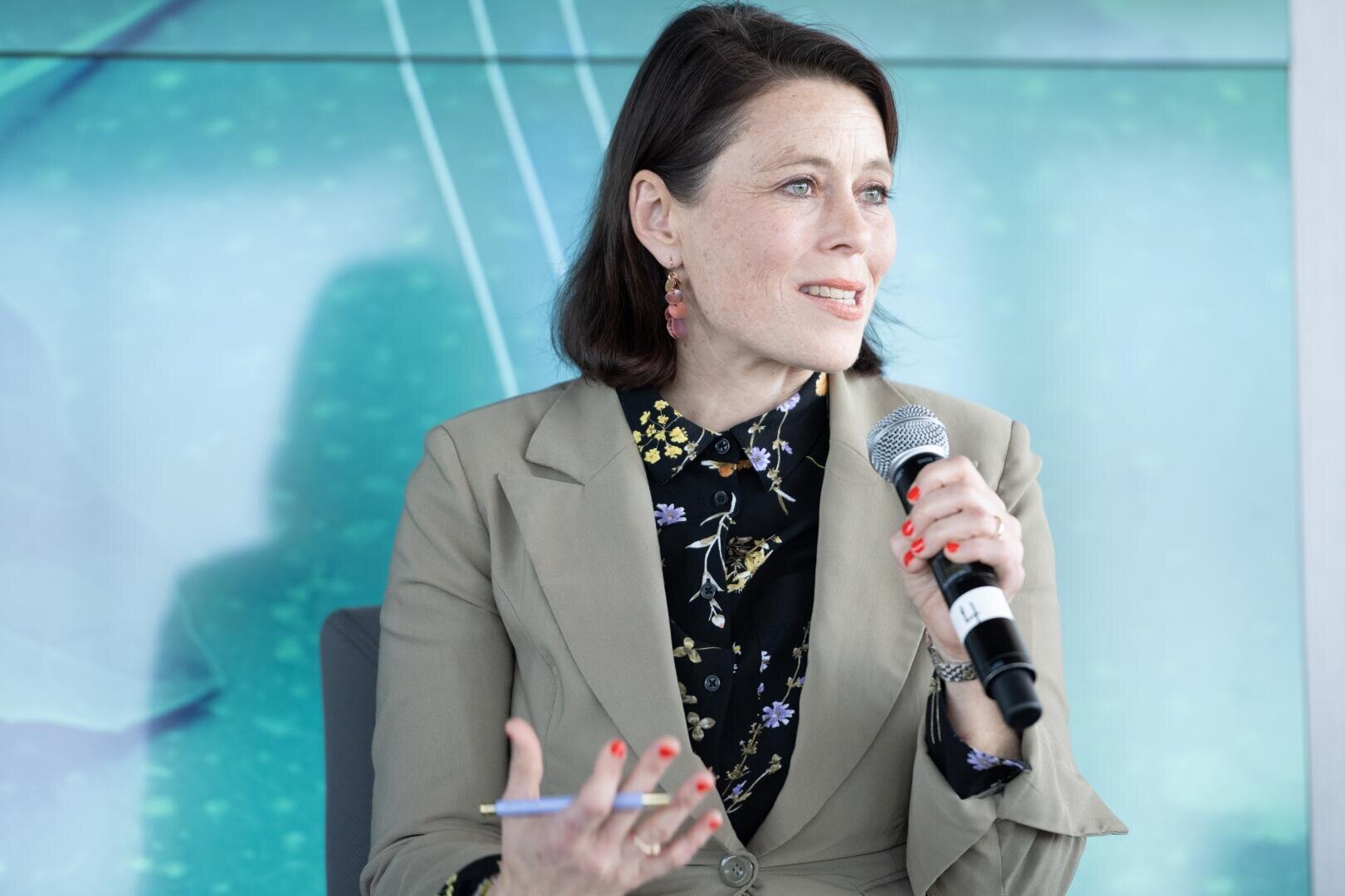 Eine Frau mit kurzen braunen Haaren, die einen beigen Blazer und ein geblümtes Hemd trägt, hält ein Mikrofon und einen Stift in der Hand und spricht bei einer Veranstaltung vor einem blaugrünen Hintergrund.