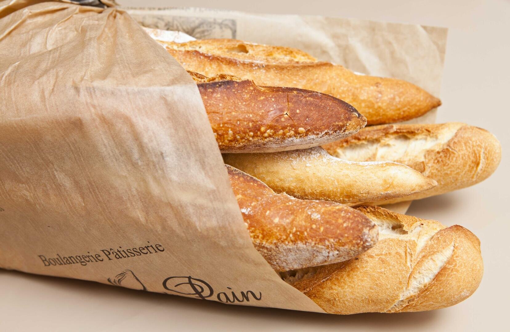 Drei frische Baguettes, teilweise in braunes Papier mit der Aufschrift „Boulangerie Pâtisserie“ eingewickelt, mit knusprigen, goldbraunen Oberflächen und einem hellbeigen Hintergrund.