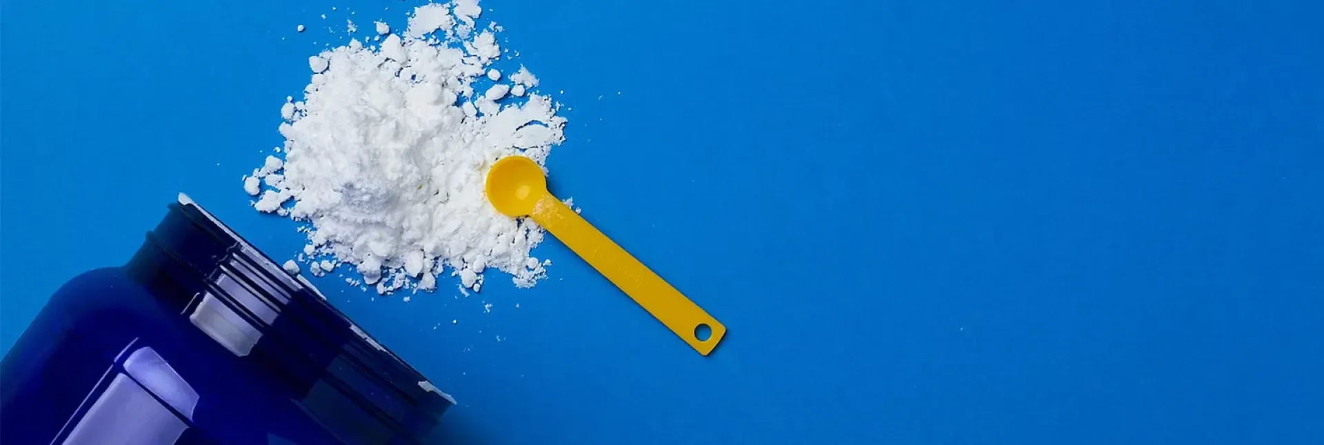 Ein blauer Behälter kippte um, wobei weißes Pulver und eine gelbe Plastikschaufel auf eine leuchtend blaue Fläche verschüttet wurden.