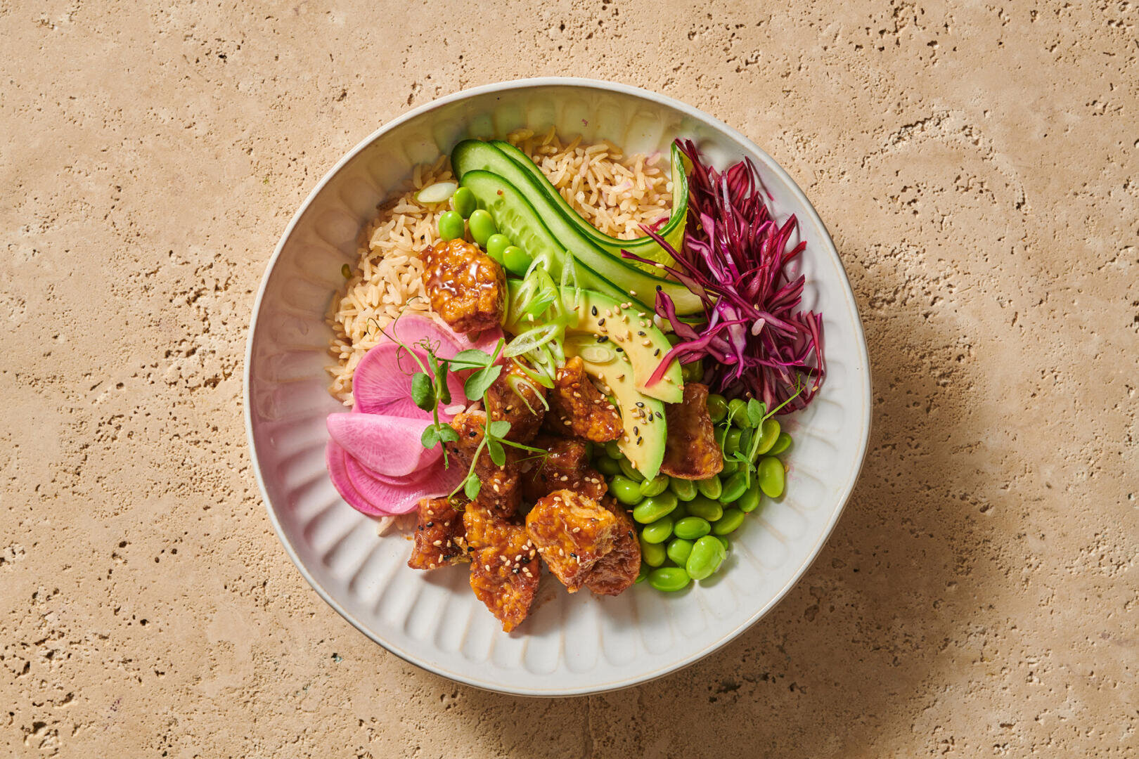 Eine Reisschüssel mit glasiertem Tofu, Avocadoscheiben, Gurke, Edamame, eingelegtem Rettich und Rotkohlstreifen.