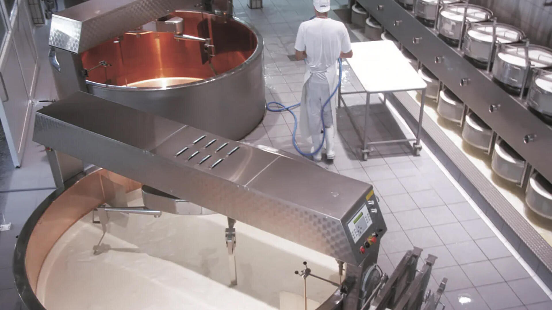 Ein Mitarbeiter in weißer Arbeitskleidung reinigt den Boden in einer Käseproduktionsstätte, in der große Metallbehälter mit Milch und Regale mit Käselaiben zu sehen sind.