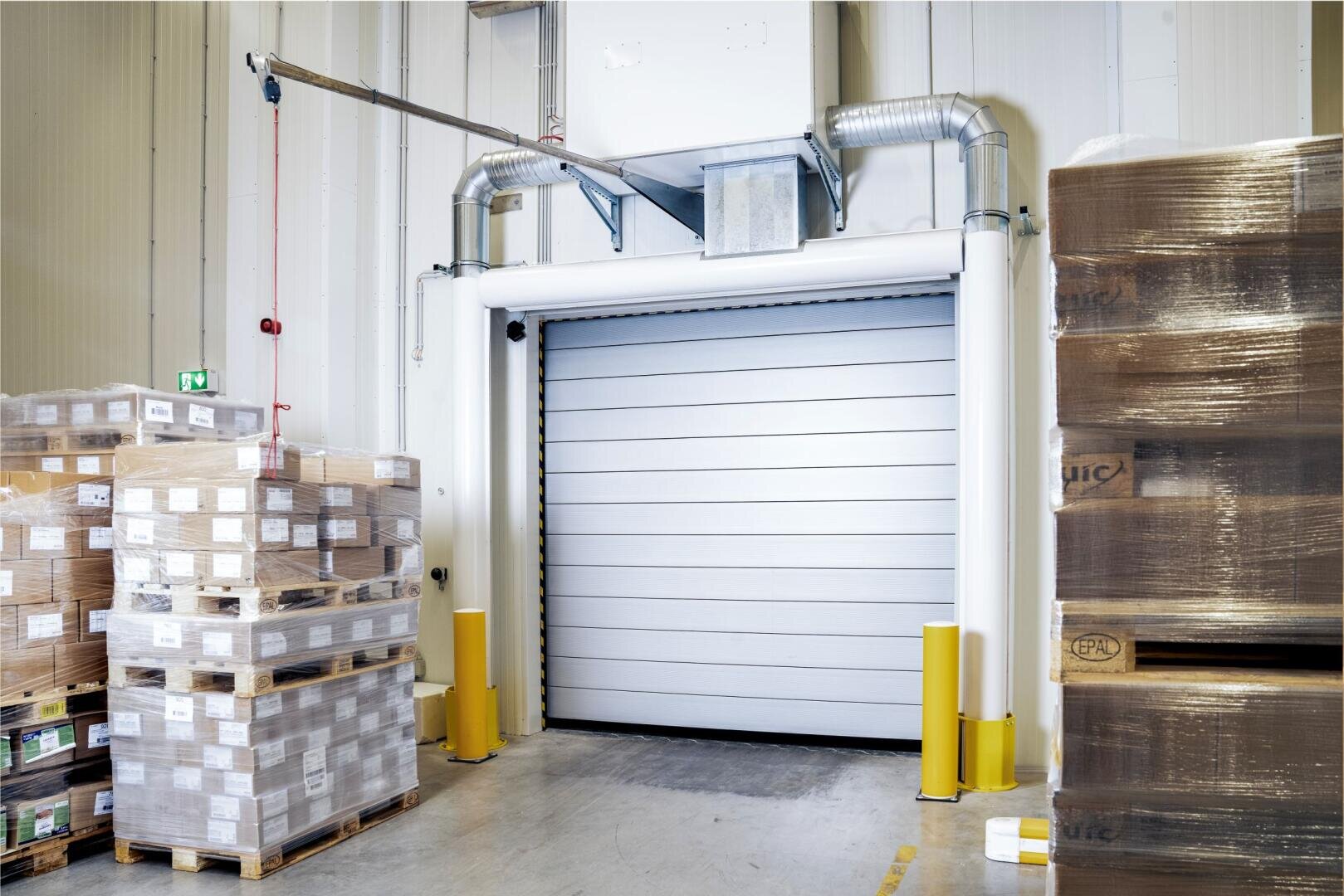 Der Innenraum eines Lagers mit einem großen, geschlossenen Rolltor, umgeben von gestapelten Paletten mit verpackten Waren und in Plastikfolie eingewickelten Gegenständen. Gelbe Sicherheitspfosten sichern den Eingang.