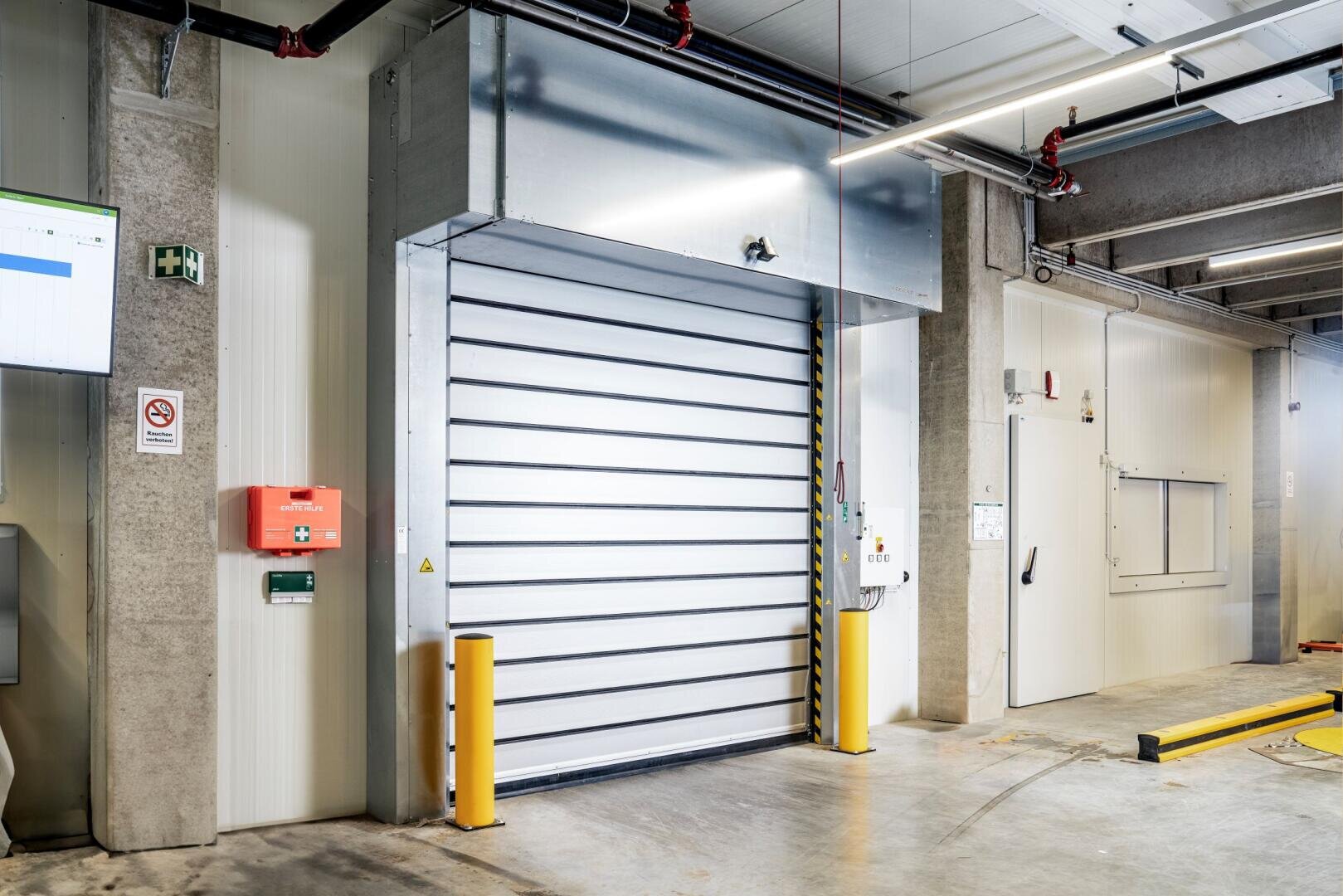 Eine moderne industrielle Laderampe mit einem großen Metallrolltor, Sicherheitspoller, Notfallknöpfen und einer weißen Wand mit einer kleinen Zugangstür – alles in einer sauberen, gut beleuchteten Umgebung.