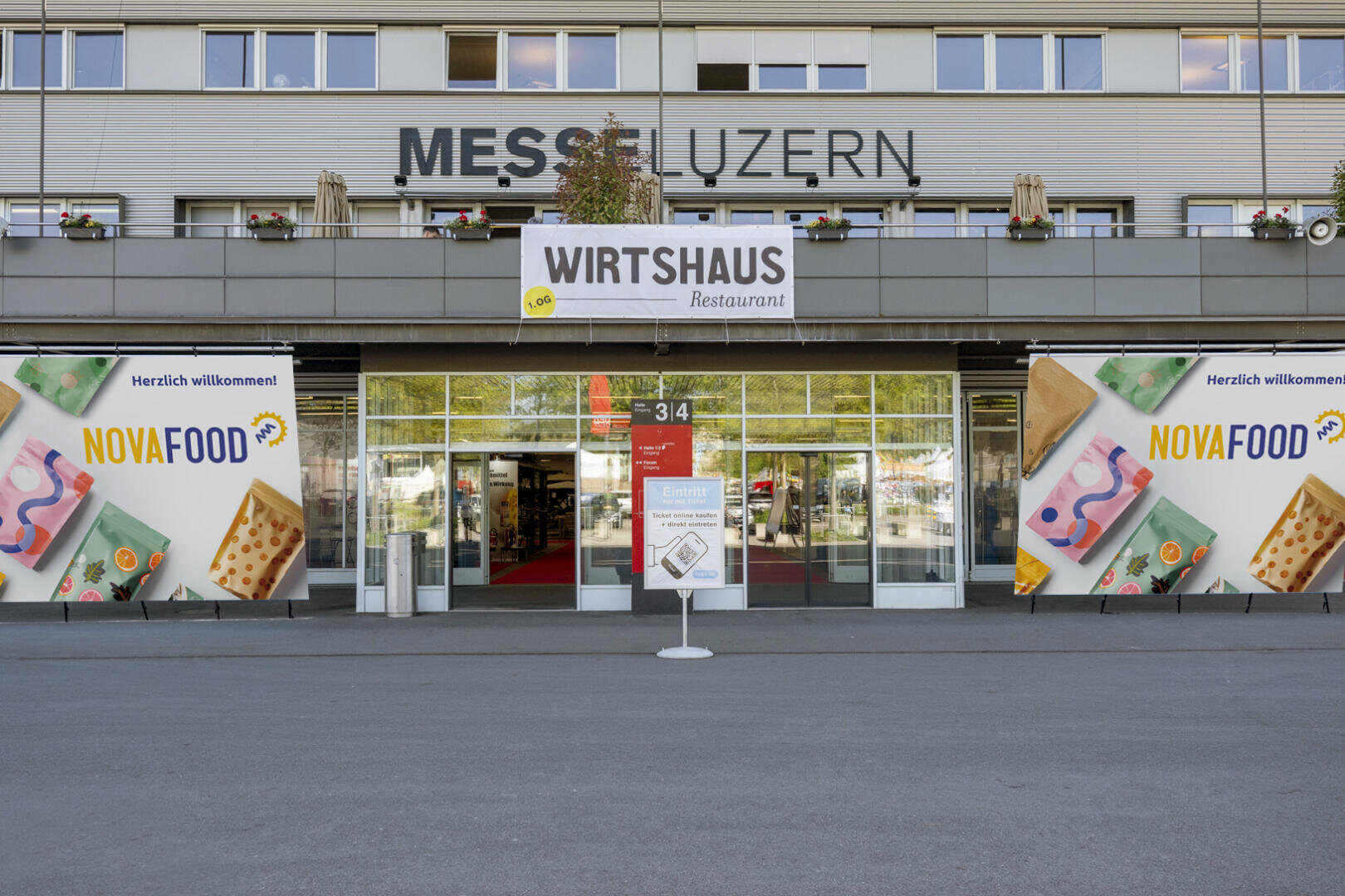 Eingang zur Messe Luzern mit großem WIRTSHAUS-Restaurant-Banner, NOVAFOOD-Plakaten auf beiden Seiten und Glastüren, in denen sich Bäume spiegeln. Vor dem Eingang steht ein Schild.