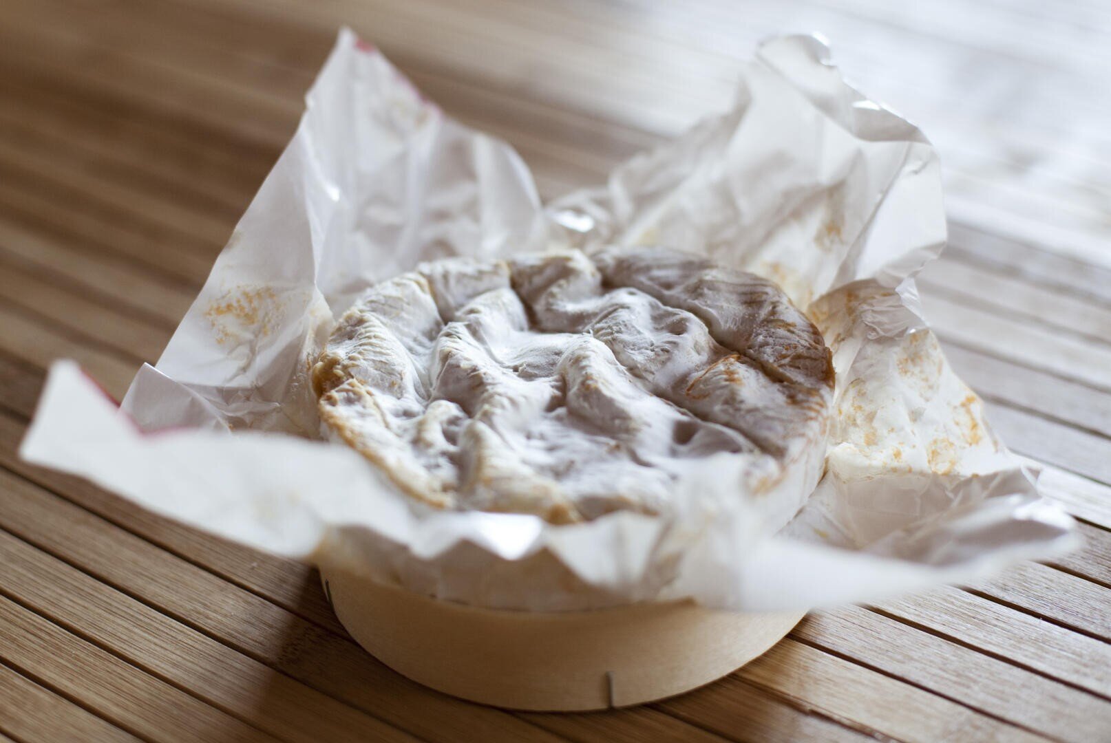 Ein runder Weichkäse mit welliger, weißer Rinde liegt, teilweise in weißes Papier eingewickelt, auf einer Holzunterlage.