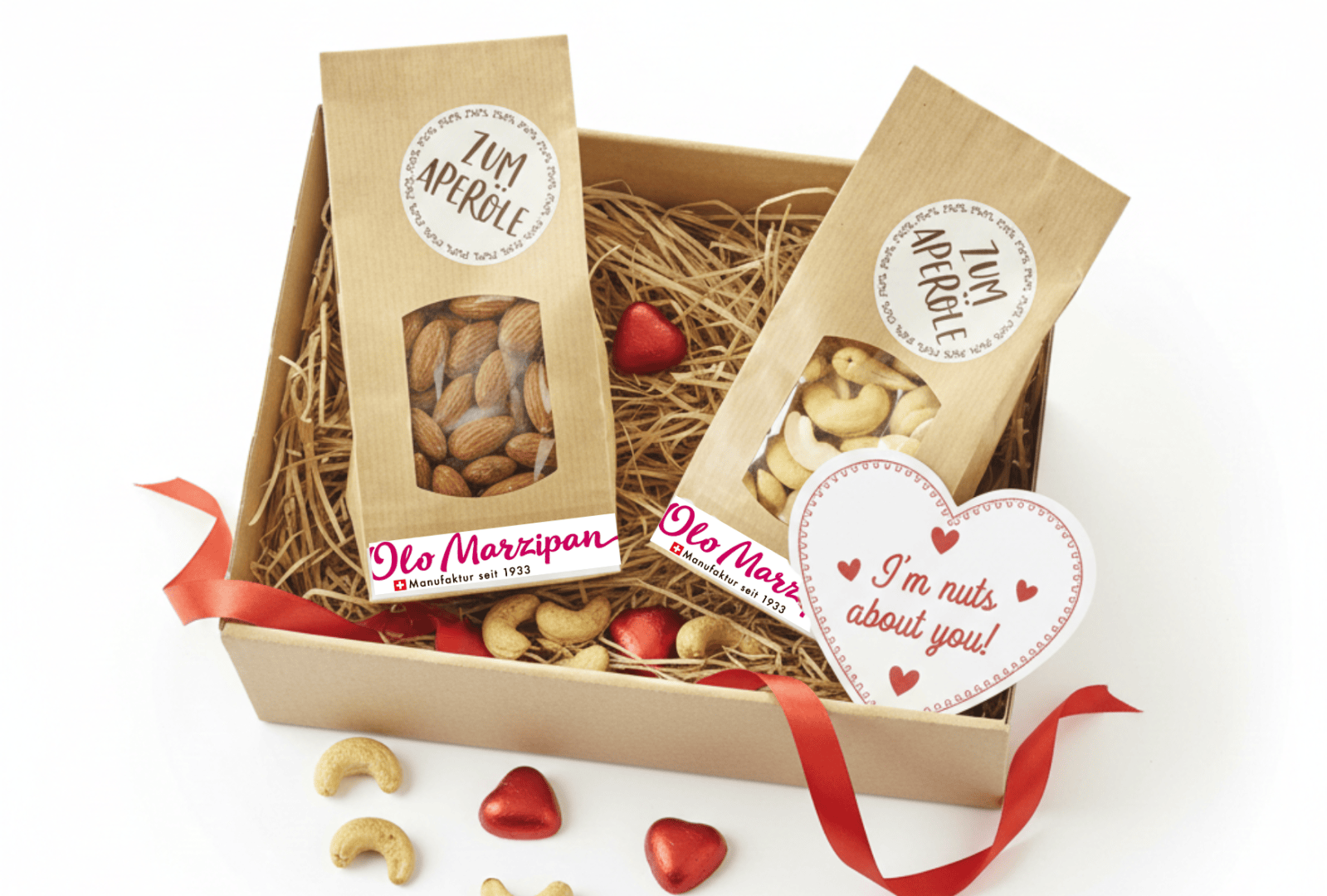Eine Geschenkbox mit Päckchen Mandeln und Cashewnüssen, roten Herzpralinen in Folie, einer roten Schleife und einer herzförmigen Karte mit der Aufschrift „Ich bin verrückt nach dir!“ auf Strohfüllung.