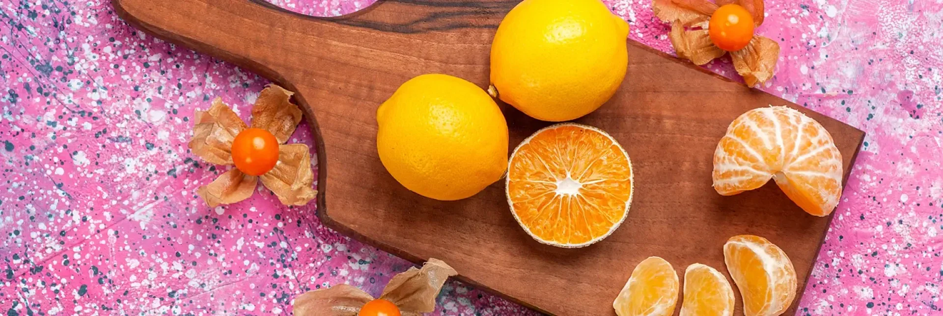 Zwei ganze Zitronen, eine halbierte Orange, geschälte Orangenscheiben und Goldbeeren liegen auf einem Holzschneidebrett, das auf einer mit rosa Spritzern verzierten Oberfläche steht.