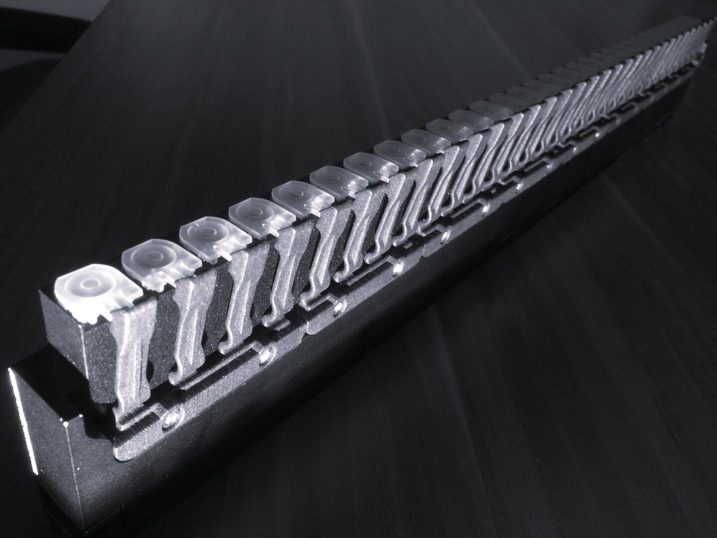 Eine lange, metallische Reihe gleichförmiger mechanischer Tasten, die vermutlich von einer Tastatur oder einer Schreibmaschine stammen, ist diagonal auf einer dunklen Fläche angeordnet.