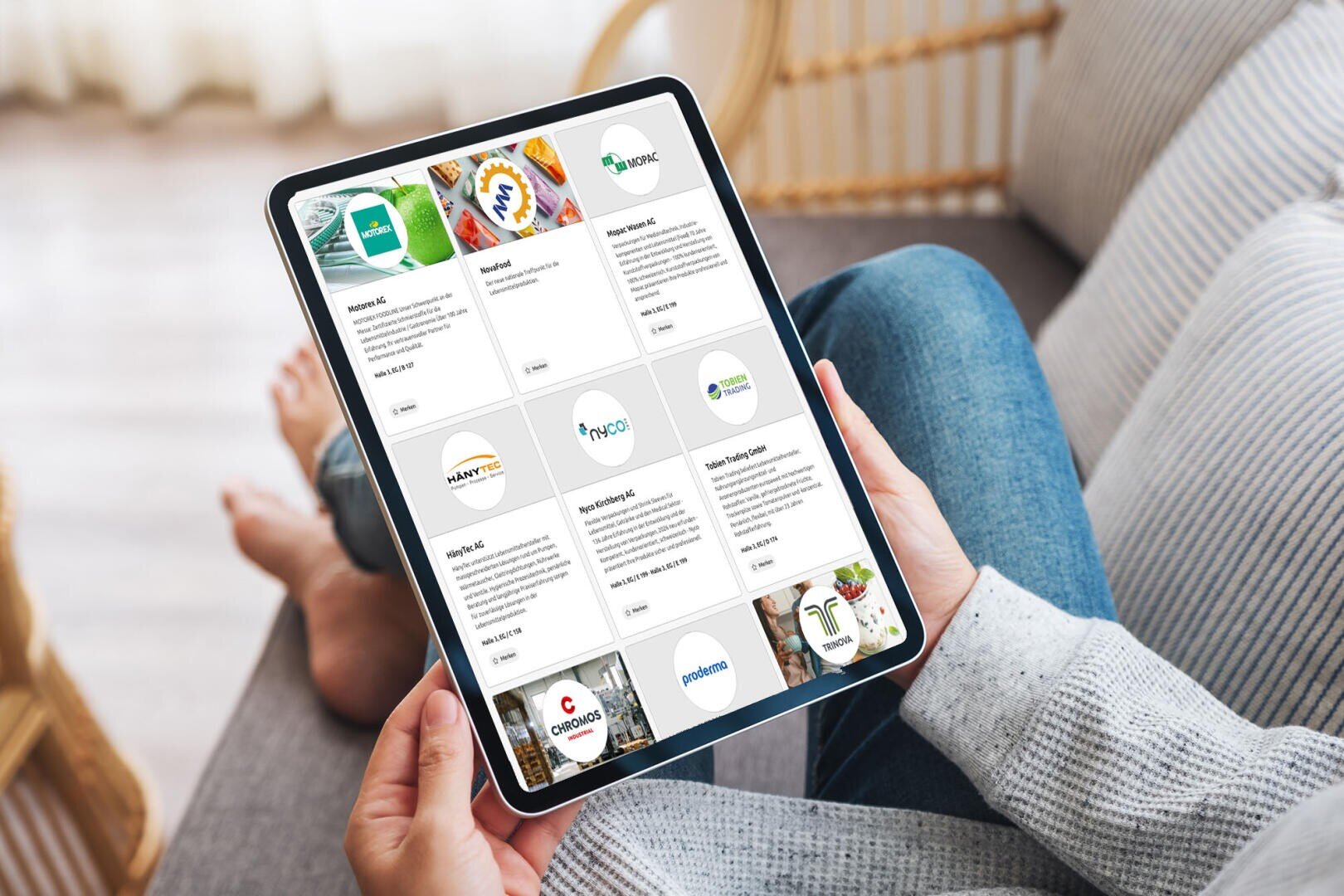 Eine Person sitzt auf einem Sofa und hält ein Tablet in der Hand, auf dem eine Tabelle mit Firmenlogos und Firmennamen zu sehen ist, was auf ein Verzeichnis oder einen Auswahlbildschirm hindeutet. Die Atmosphäre wirkt ungezwungen und entspannt.