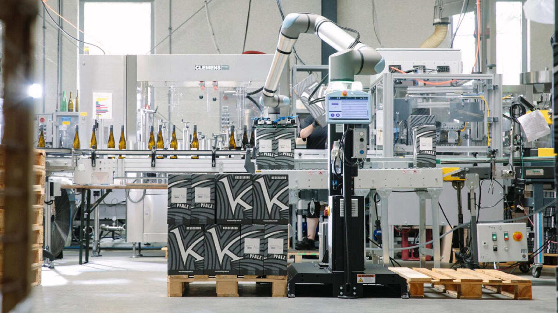 In einer modernen Fabrikumgebung verpackt ein Roboterarm Weinflaschen in Kartons. Im Vordergrund sind gestapelte Kartons und leere Paletten zu sehen, im Hintergrund Maschinen und Flaschen auf einem Förderband.
