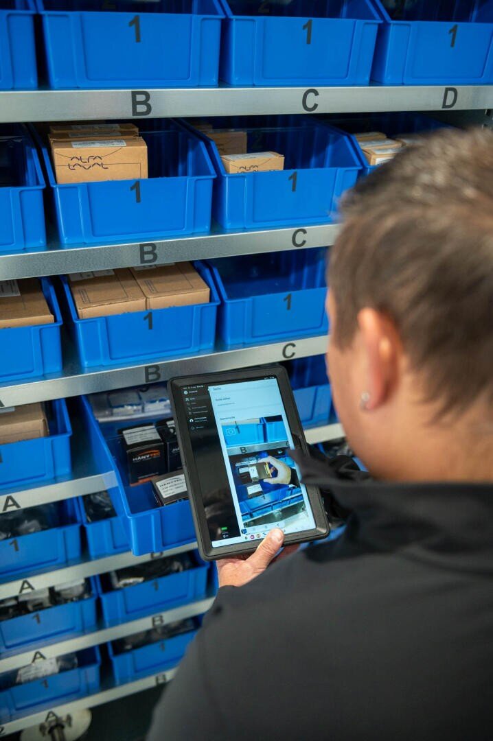 Eine Person nutzt ein Tablet, um den Bestand in blauen, mit Buchstaben und Zahlen beschrifteten Lagerbehältern in den Regalen zu scannen oder zu überprüfen.