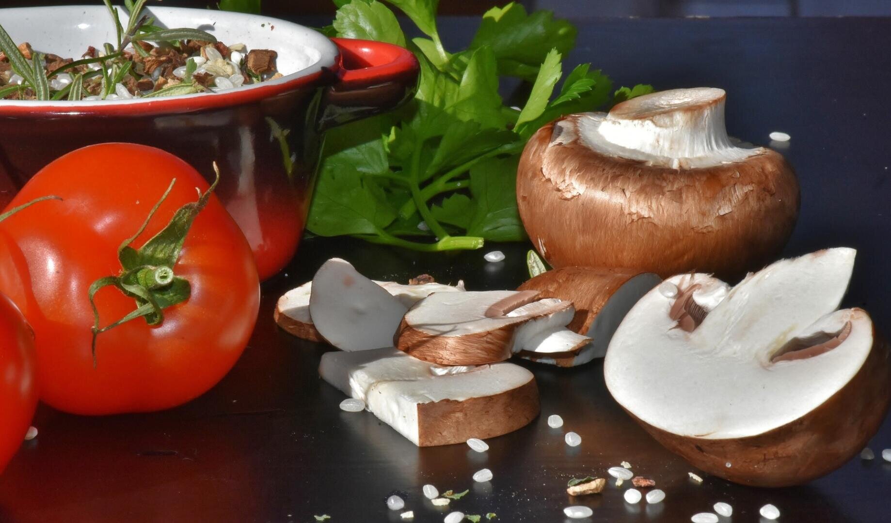 Eine rote Tomate, frische Petersilie und braune Champignons – teils ganz, teils in Scheiben geschnitten – auf einer dunklen Unterlage neben einem kleinen Topf mit Kräutern angeordnet.