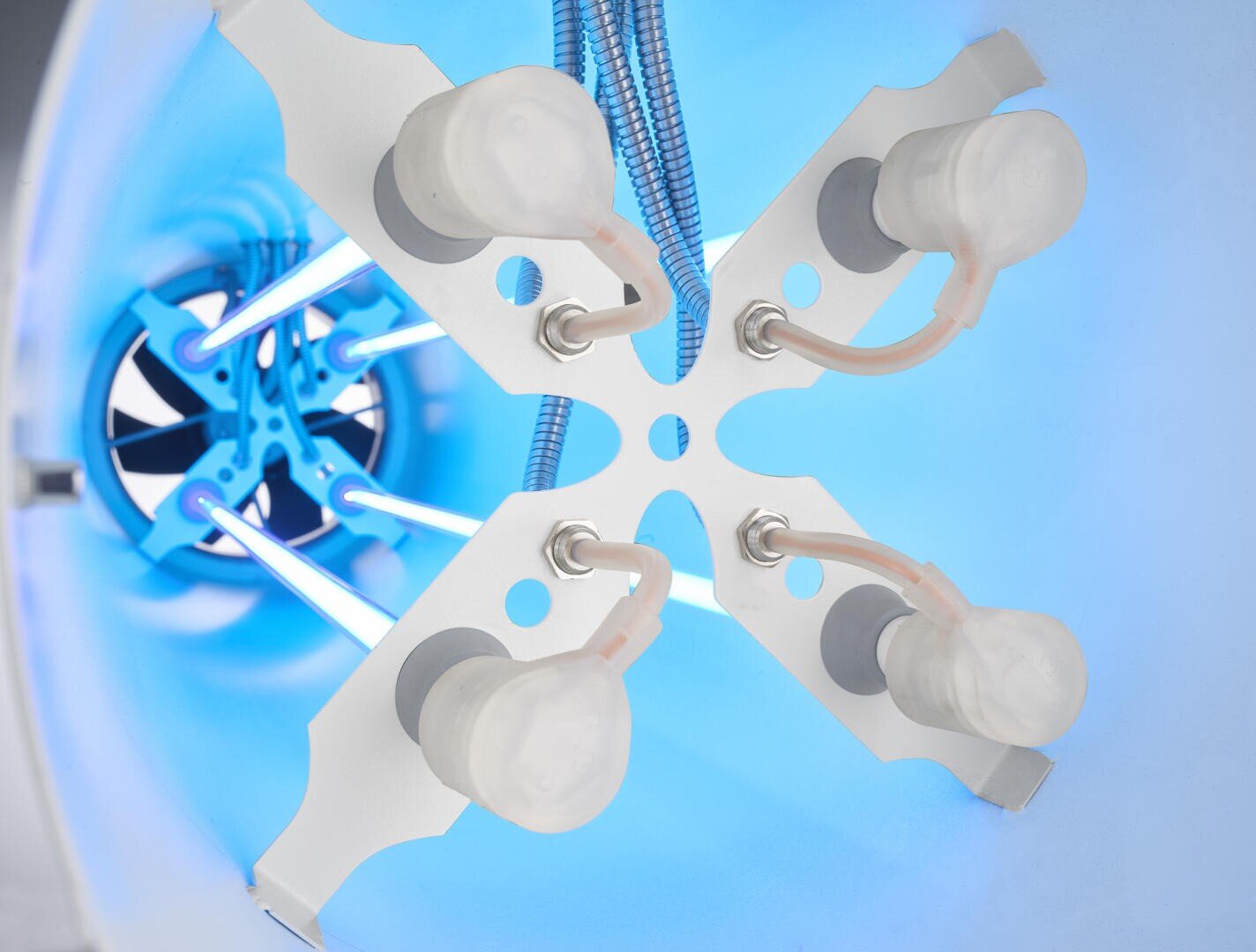 Nahaufnahme eines Laborgeräts mit vier durchsichtigen Behältern, die an Schläuchen befestigt sind und auf einer weißen, X-förmigen Halterung montiert sind, mit blauer Beleuchtung und Metallstangen im Hintergrund.