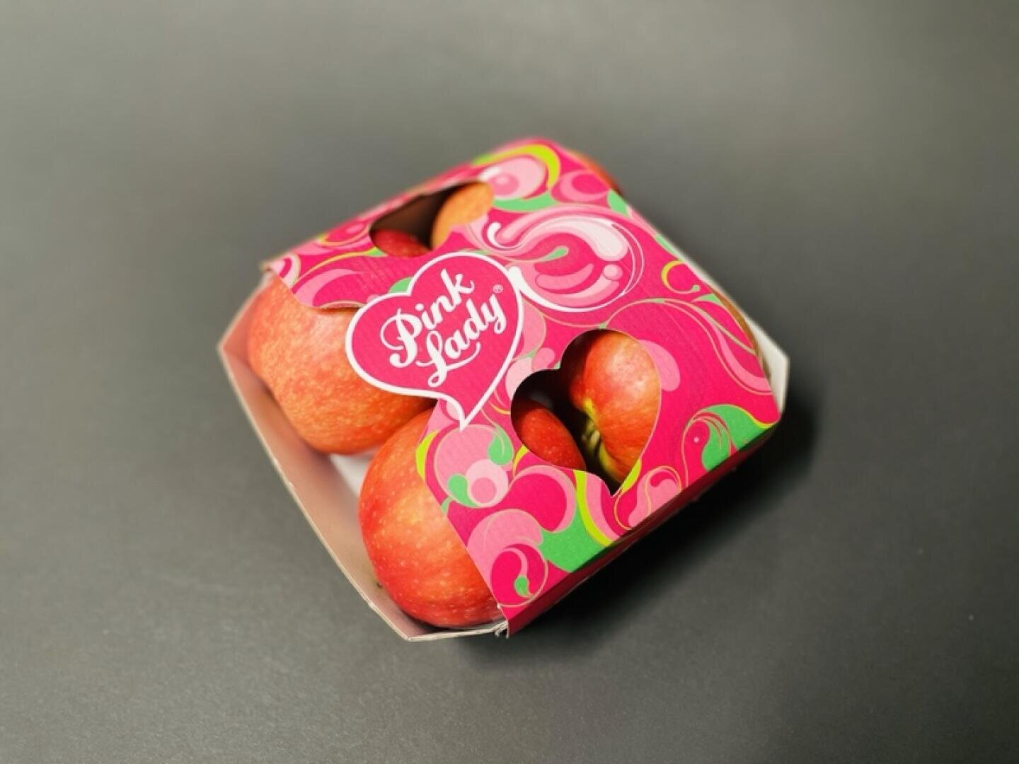 Auf einer dunklen Fläche steht eine Pappschale mit Pink-Lady-Äpfeln, die teilweise von einer bunten rosa-grünen Verpackung mit herzförmigen Ausschnitten bedeckt ist.