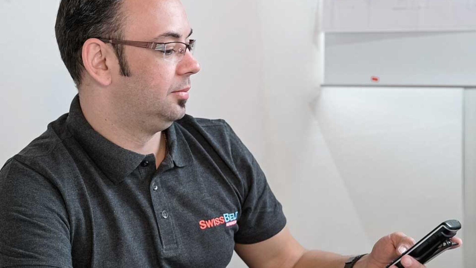 Ein Mann mit Brille und einem dunklen Polohemd mit Swissbau-Logo sitzt in einem Raum und schaut auf sein Smartphone, im Hintergrund ist eine Whiteboard-Tafel zu sehen.
