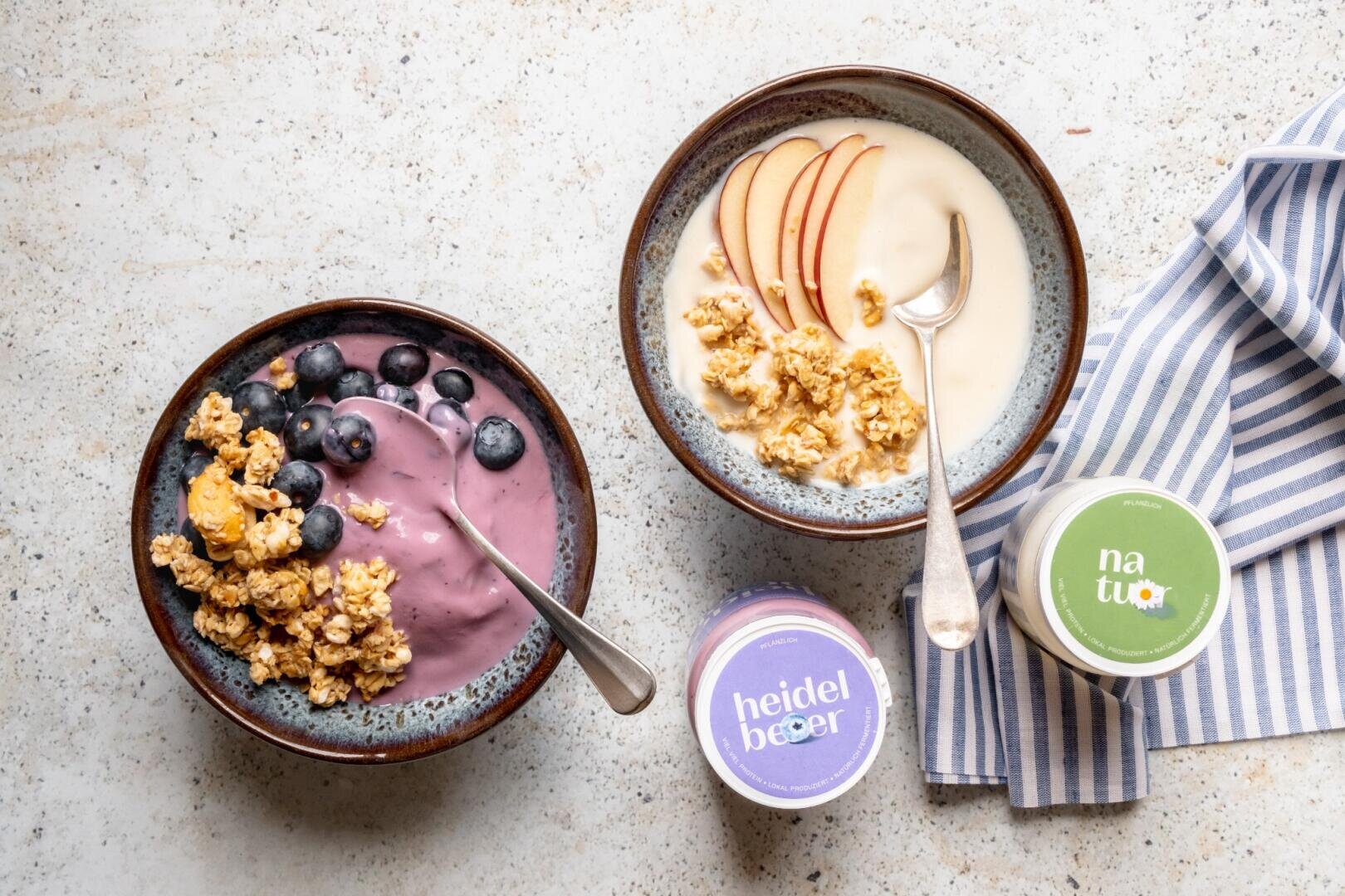 Zwei Schalen Joghurt, garniert mit Müsli, Heidelbeeren und Apfelscheiben, daneben zwei Becher Joghurt auf einer blau-weiß gestreiften Serviette.
