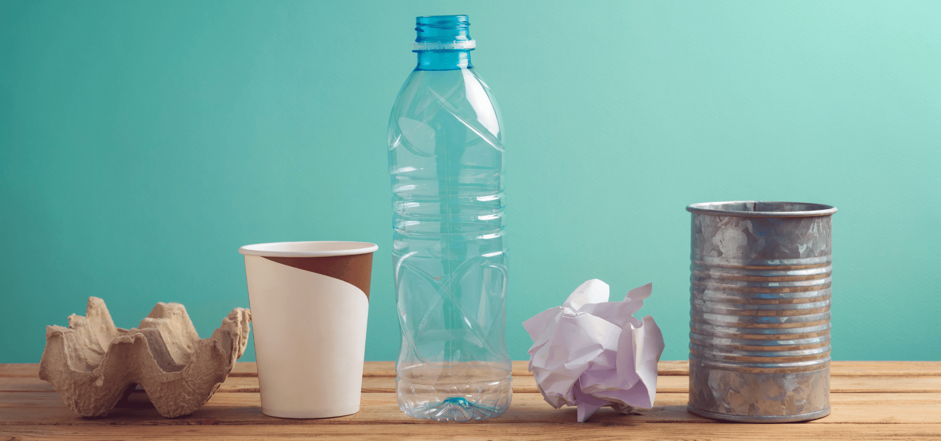 Auf einer Holzunterlage vor einem blaugrünen Hintergrund angeordnete Recyclinggegenstände: ein Eierkarton aus Pappe, ein Kaffeebecher aus Papier, eine leere, durchsichtige Plastikwasserflasche, ein zerknülltes Stück weißes Papier und eine leere Metalldose.