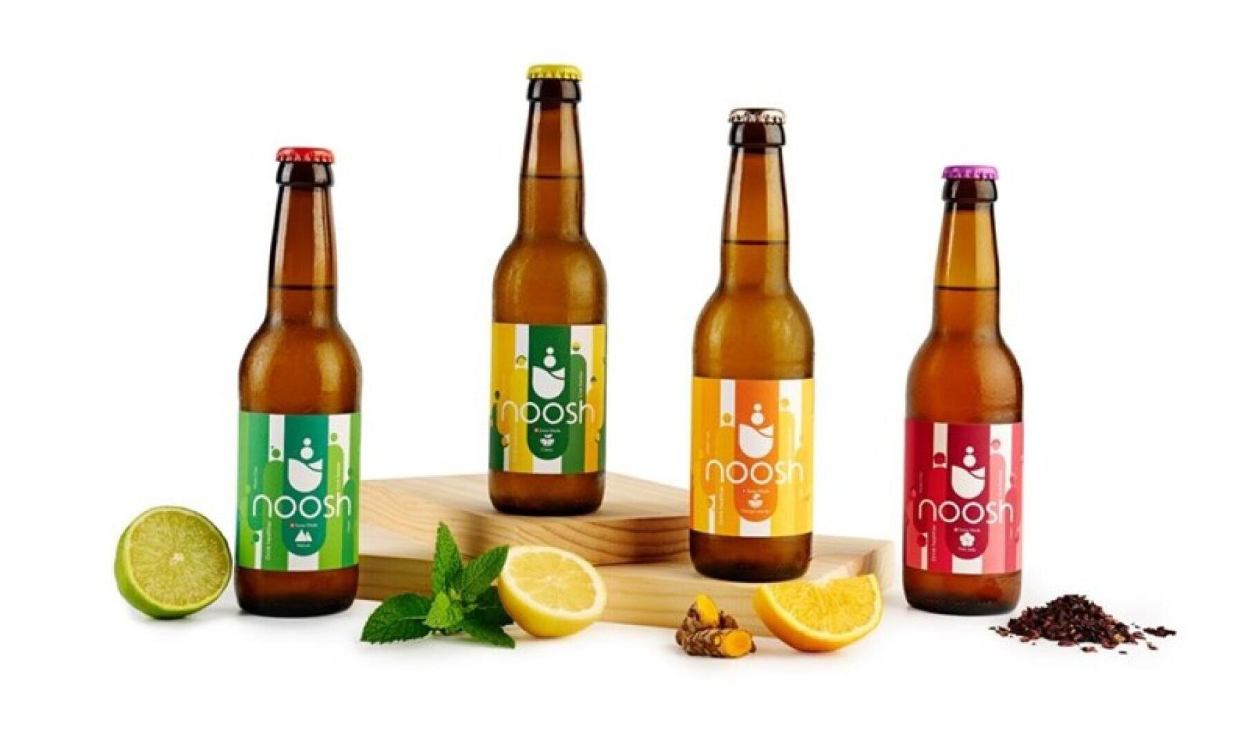 Vier bernsteinfarbene Glasflaschen mit dem Getränk Noosh und bunten Etiketten, präsentiert zusammen mit frischen Limetten, Zitronen, Minze, Kurkuma und getrockneten Hibiskusblüten.
