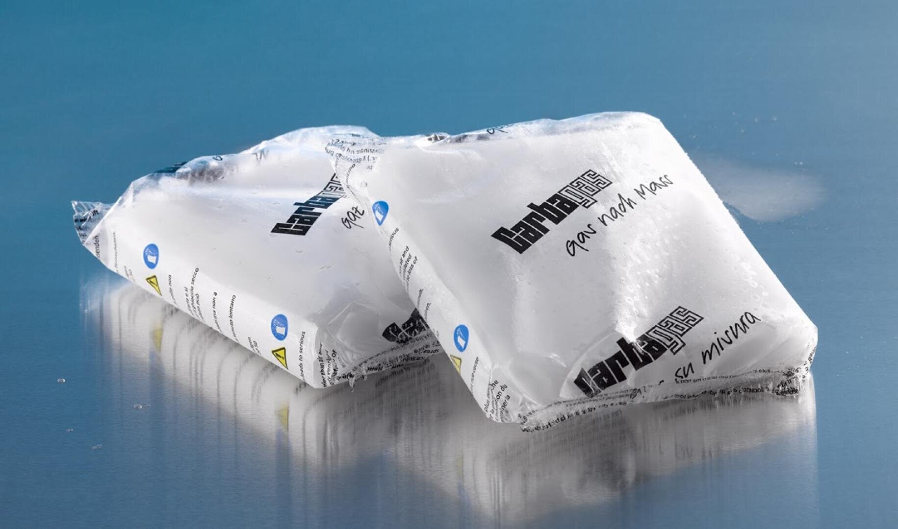 Auf einer blauen Fläche liegen zwei versiegelte Plastikverpackungen, die weißes Trockeneis enthalten. Die Verpackungen sind mit schwarzem und grauem Text beschriftet, und auf den Verpackungen sowie um sie herum ist etwas Reif zu sehen.