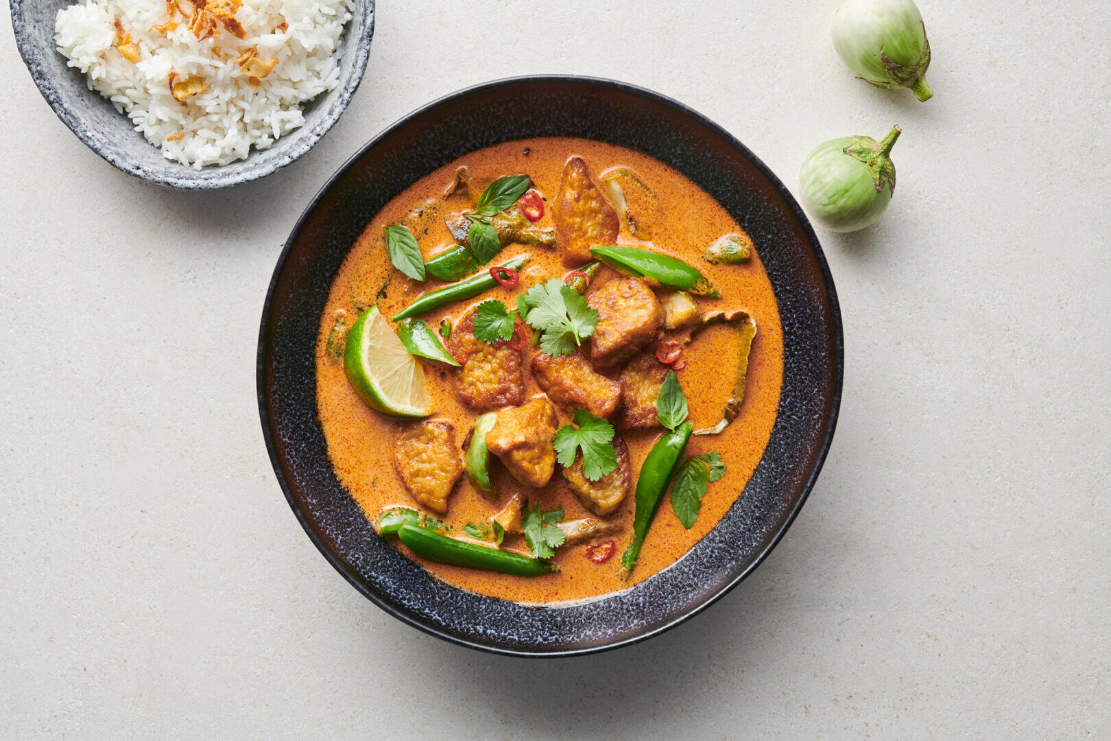 Eine Schüssel rotes Curry mit Tofu, grünen Chilischoten und Kräutern, serviert mit weißem Reis als Beilage und zwei kleinen Auberginen.