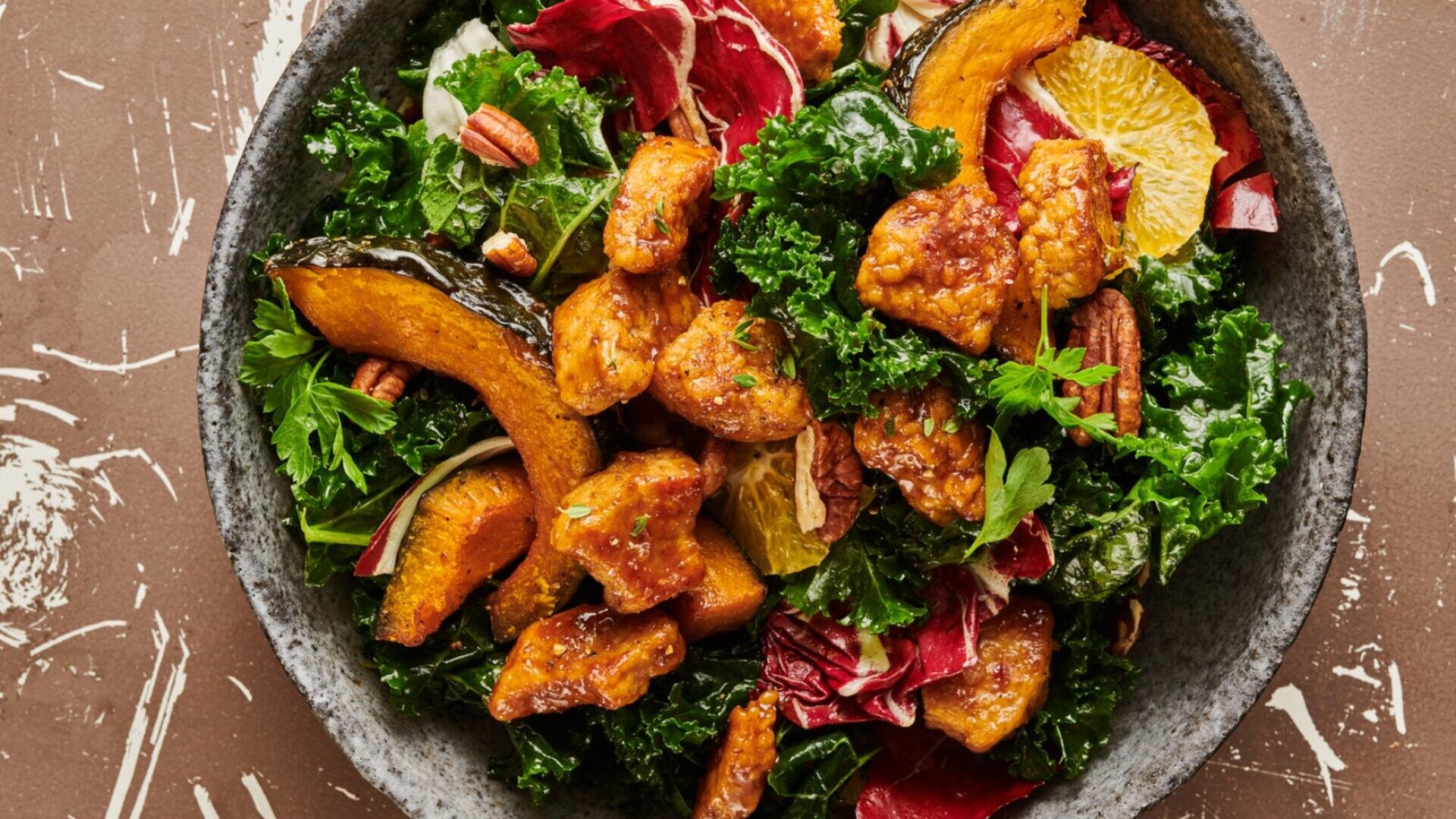 Eine Salatschüssel mit Grünkohl, geröstetem Kürbis, Pekannüssen, Orangenscheiben und glasierten Proteinstückchen.