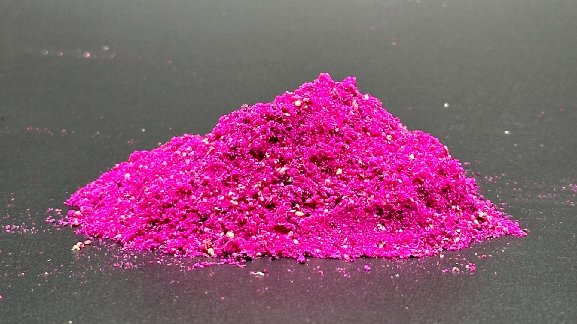 Auf einer schlichten, dunklen Fläche liegt ein kleiner Haufen leuchtend magentafarbenen Pulvers, um den herum feine und etwas gröbere Partikel verstreut sind.