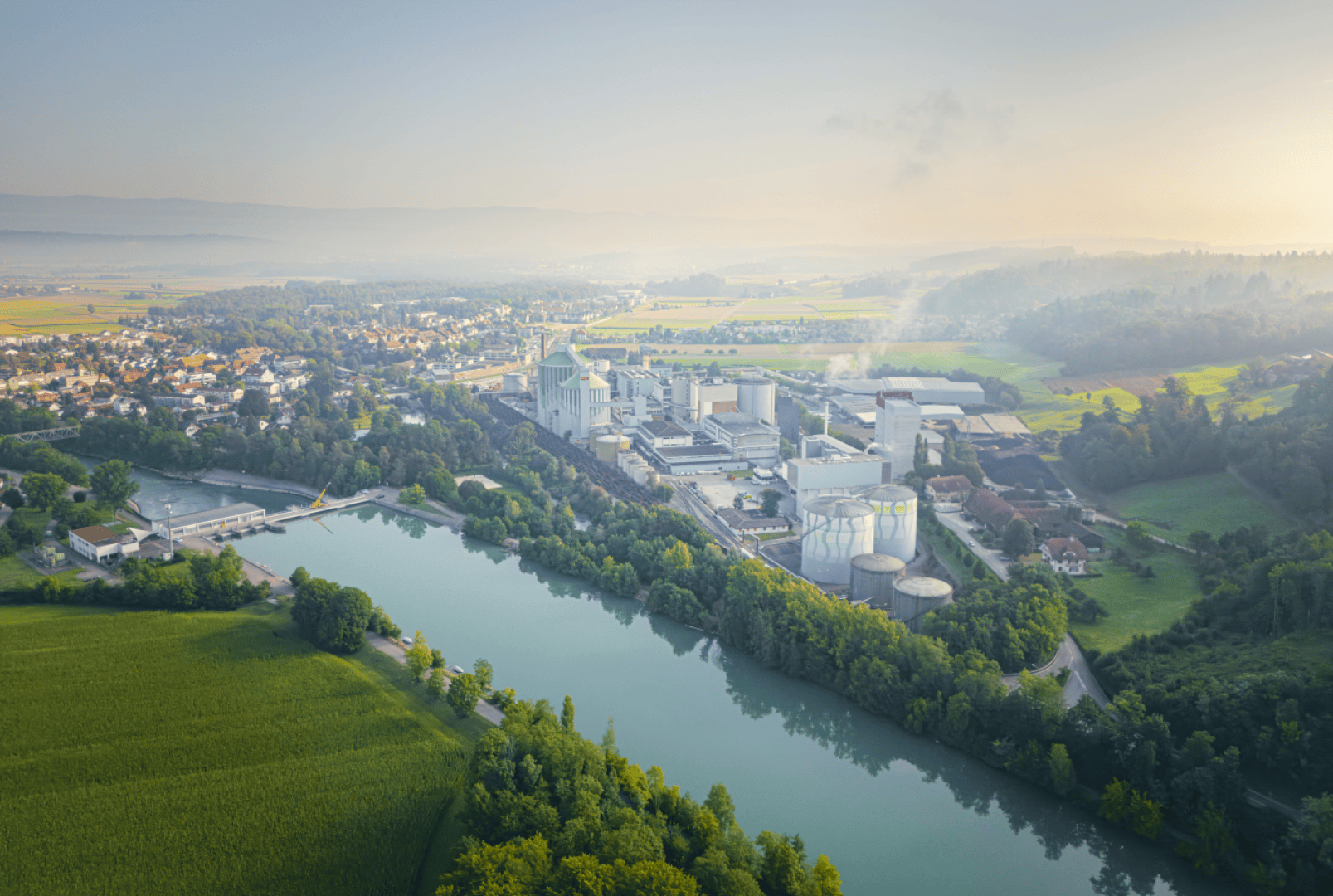 Luftaufnahme eines Industriekomplexes an einem Fluss, umgeben von grünen Feldern und Bäumen; im Hintergrund sind unter einem dunstigen Himmel eine kleine Stadt und Hügel zu sehen.