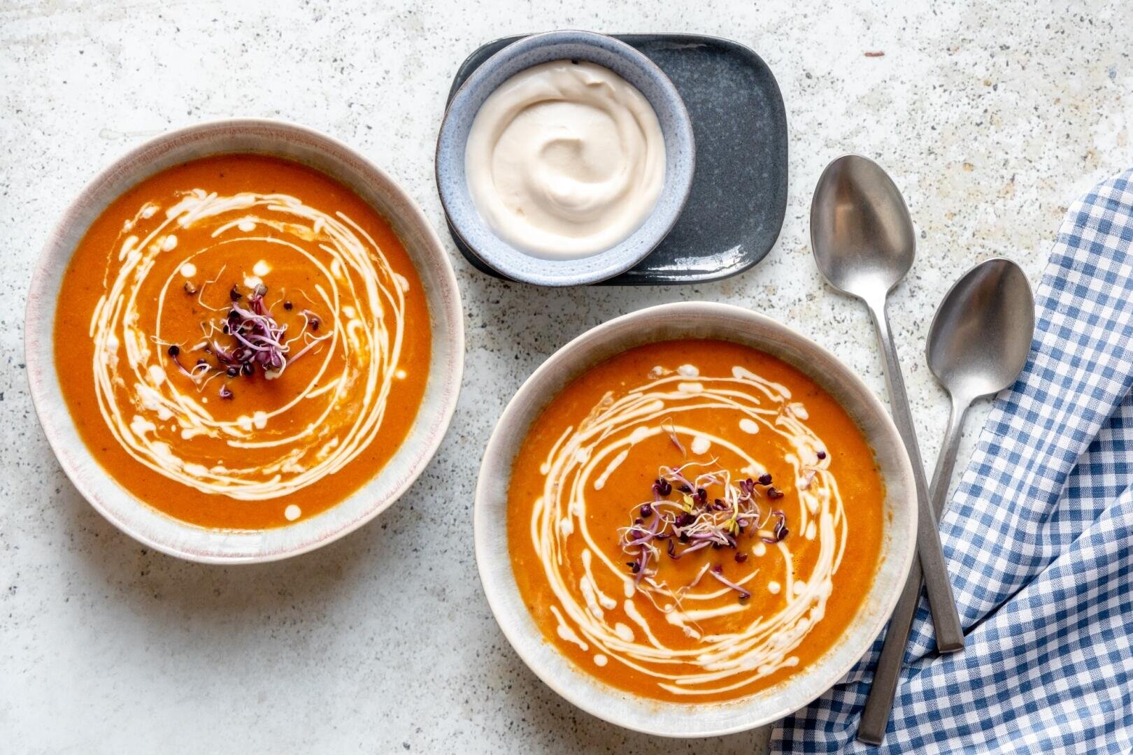 Zwei Schüsseln Tomatensuppe, mit Sahne verfeinert und mit Sprossen garniert, serviert mit einem kleinen Schälchen Sahne und zwei Löffeln auf einer karierten Serviette.