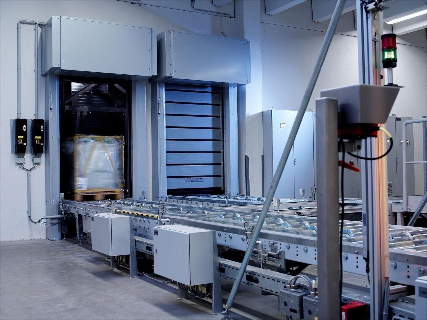 Ein modernes industrielles Fördersystem mit Rollen zum Transport von Paletten, automatischen Türen, Bedienfeldern und Sicherheitsleuchten in einer sauberen, gut beleuchteten Fabrikumgebung.