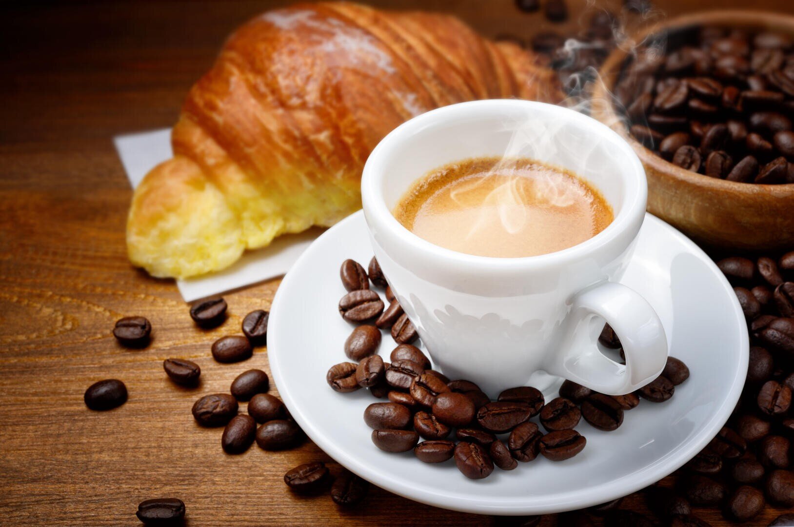 Eine dampfende Tasse Espresso auf einer Untertasse, umgeben von Kaffeebohnen, im Hintergrund ein Croissant.