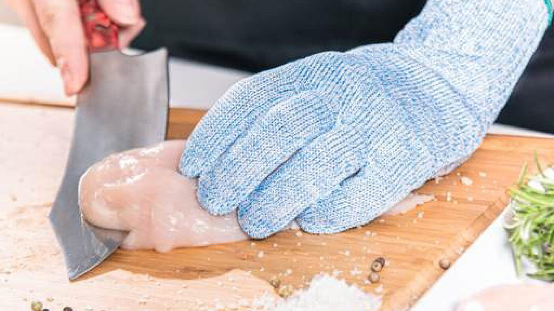 Eine Person, die einen blauen, schnittfesten Handschuh trägt, schneidet mit einem großen Messer rohe Hähnchenbrust auf einem Holzschneidebrett in Scheiben.