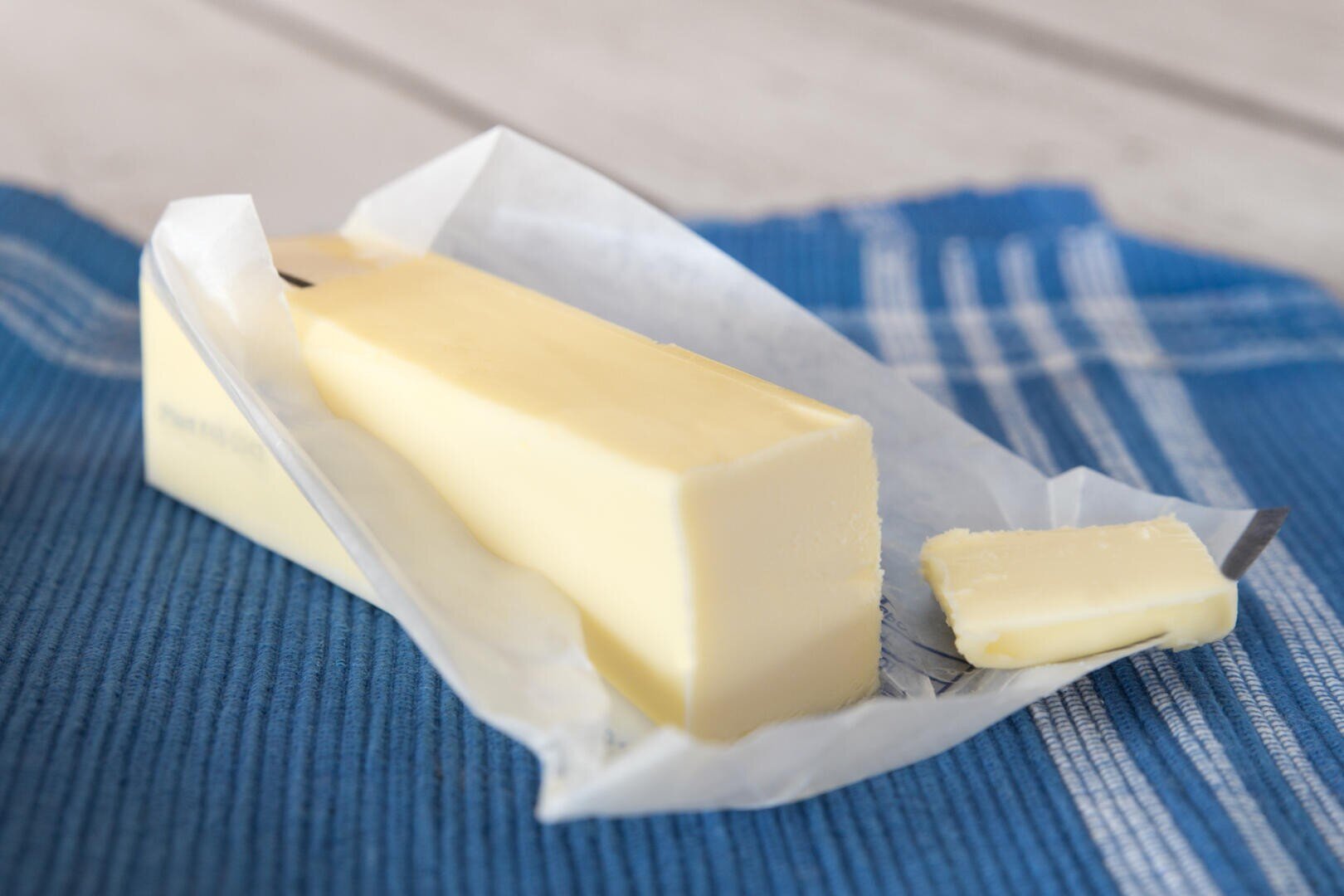 Ein Stück Butter, teilweise von der Verpackung befreit und auf einem Stück Wachspapier liegend, von dem ein kleines Stück abgeschnitten ist, auf einem blau gestreiften Tuch.