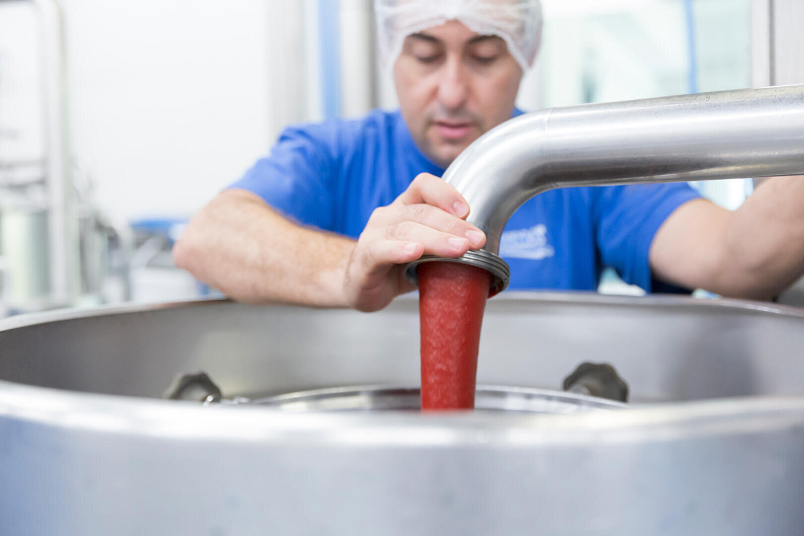 Eine Person mit Haarnetz und blauem Hemd leitet eine rote Substanz durch ein Metallrohr in einen großen Edelstahltank.