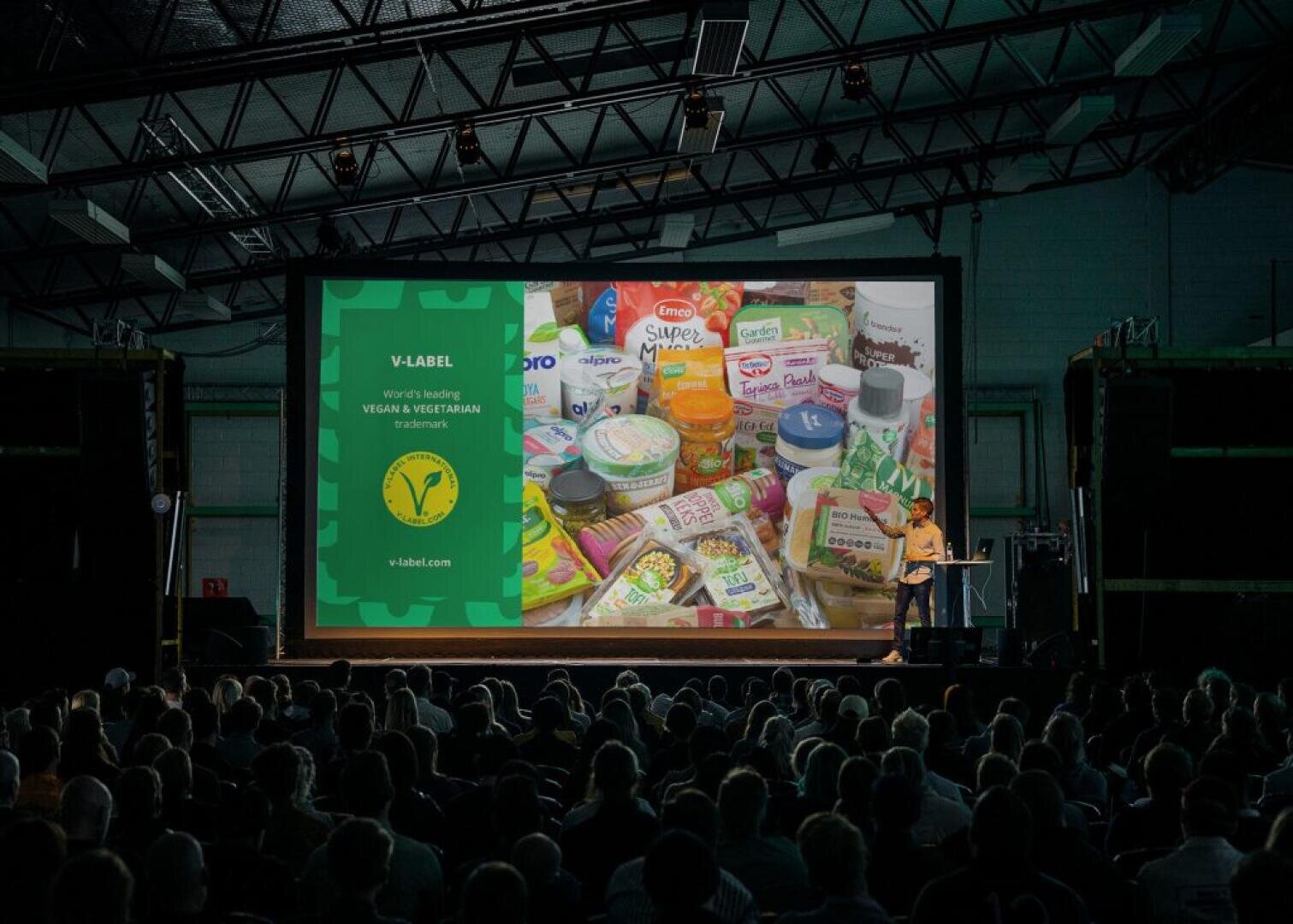 Ein Moderator spricht vor einem sitzenden Publikum in einem großen Saal und zeigt eine projizierte Folie über das V-Label sowie verschiedene vegane Produkte und Marken.