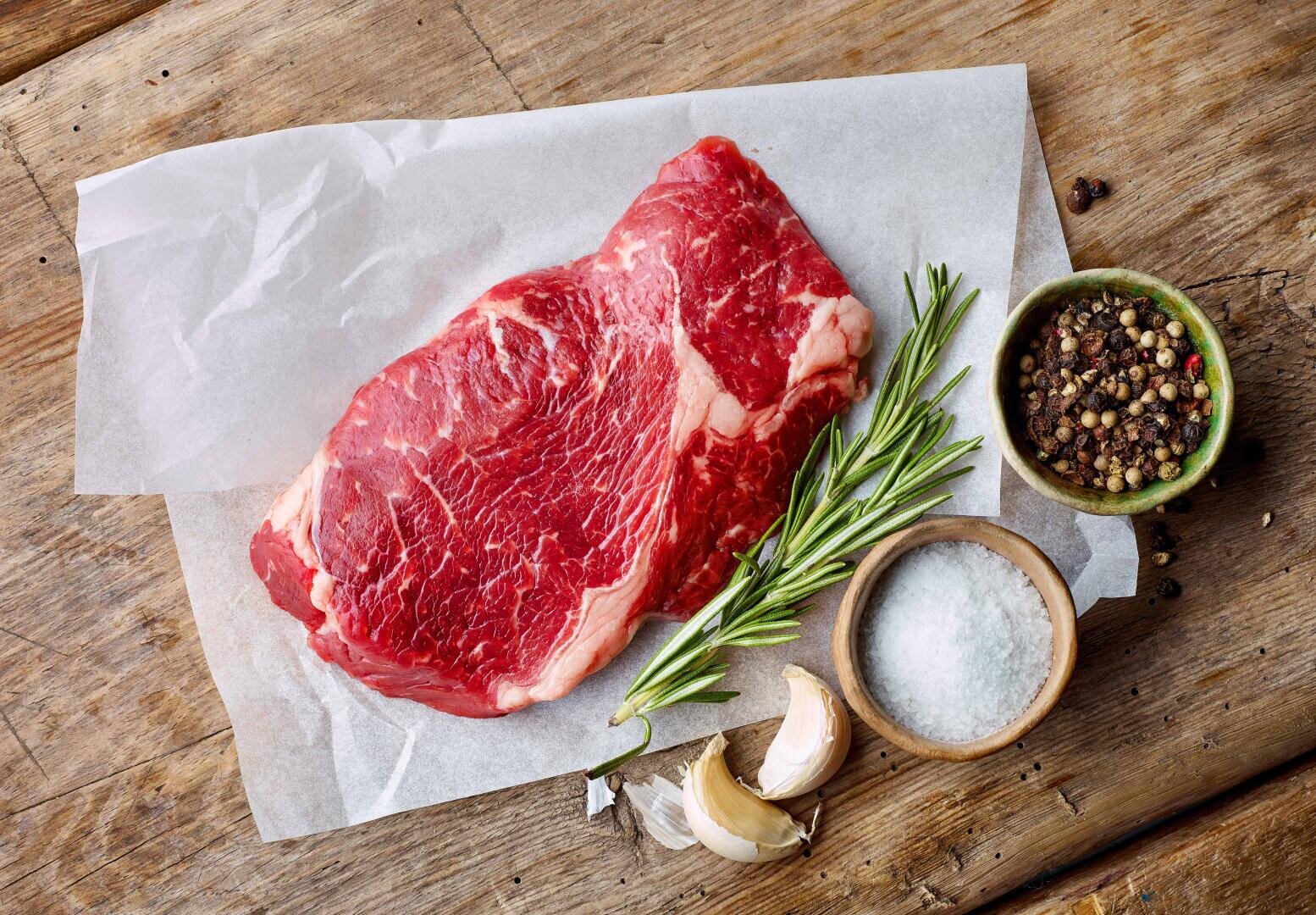Ein rohes Steak auf Backpapier, dazu Rosmarinzweige, eine Schale mit gemischten Pfefferkörnern, eine Schale mit grobem Salz und Knoblauchzehen auf einer Holzunterlage.