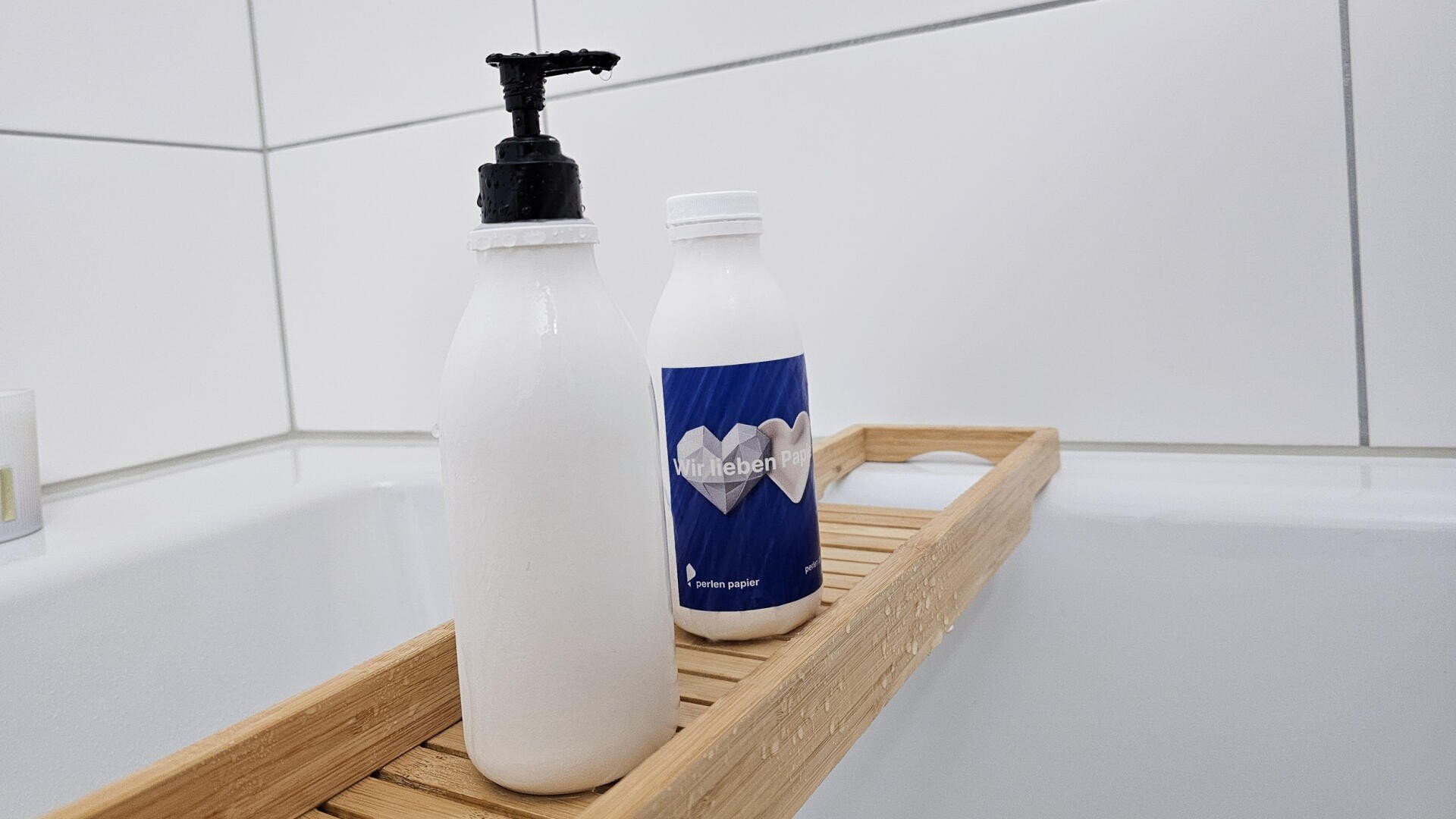 Zwei weiße Flaschen, eine mit einer Pumpe und eine mit einem blauen Etikett, stehen auf einem hölzernen Badetablett über einer weißen Badewanne, hinter der sich geflieste Wände befinden.