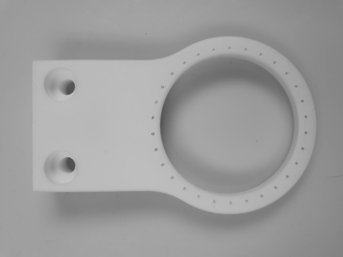 Weiße Kunststoffhalterung mit zwei großen Löchern an der rechteckigen Basis und einem kreisförmigen Ring, der an seinem Rand kleine, gleichmäßig verteilte Löcher aufweist, fotografiert vor einem einfarbigen Hintergrund.