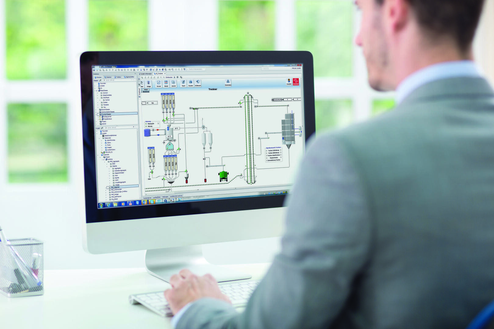 Ein Mann im Anzug arbeitet an einem Schreibtisch an einem Computer, auf dessen Bildschirm eine technische Software-Oberfläche mit Prozessablaufdiagrammen zu sehen ist, in einem hellen Büro mit großen Fenstern im Hintergrund.