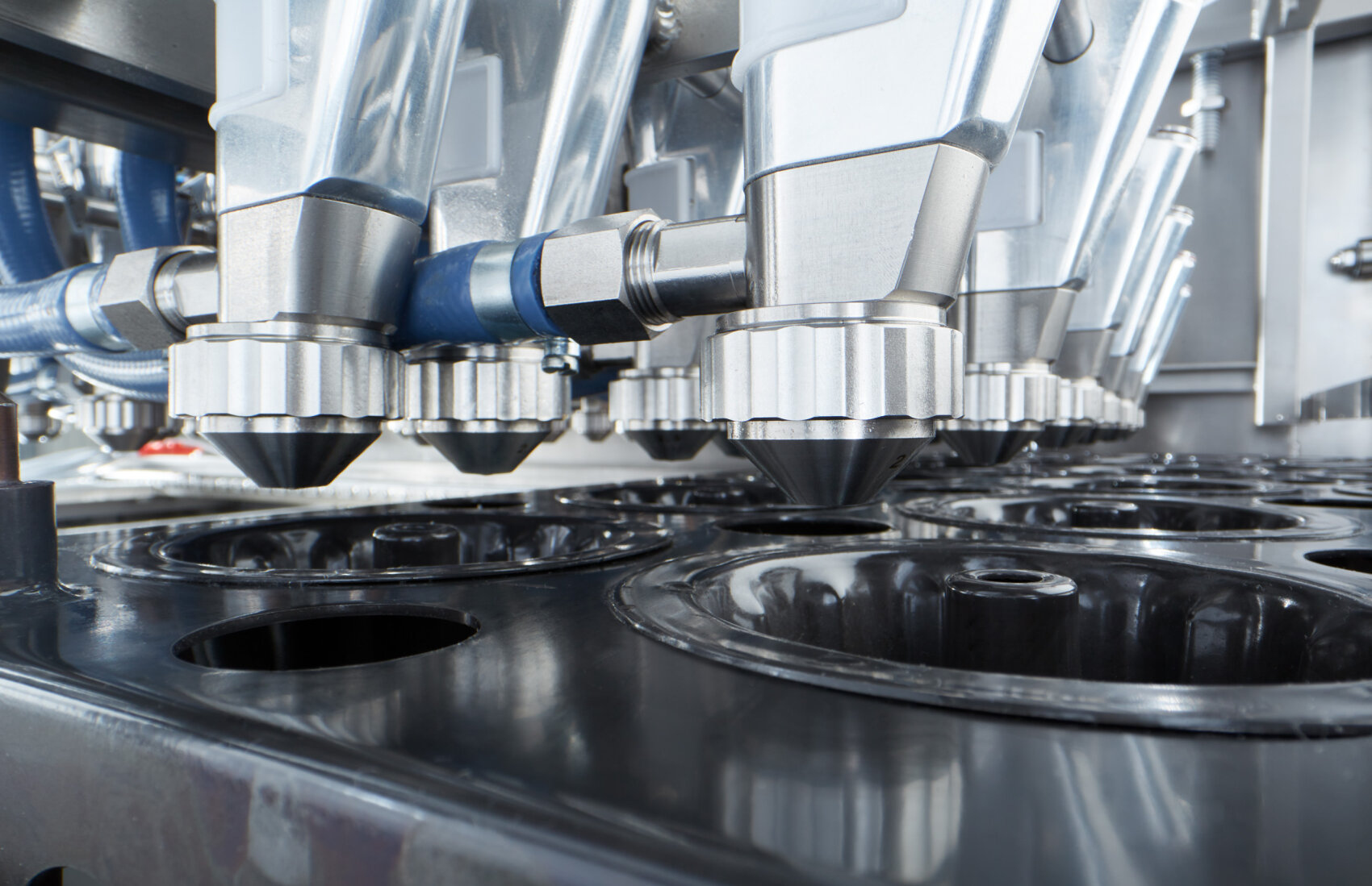 Nahaufnahme einer automatisierten Industriemaschine mit mehreren Metalldüsen, die über schwarzen, runden Formen positioniert sind; diese wird vermutlich zum Abfüllen oder zur Verarbeitung von Lebensmitteln oder flüssigen Produkten in einer Fabrikumgebung eingesetzt.